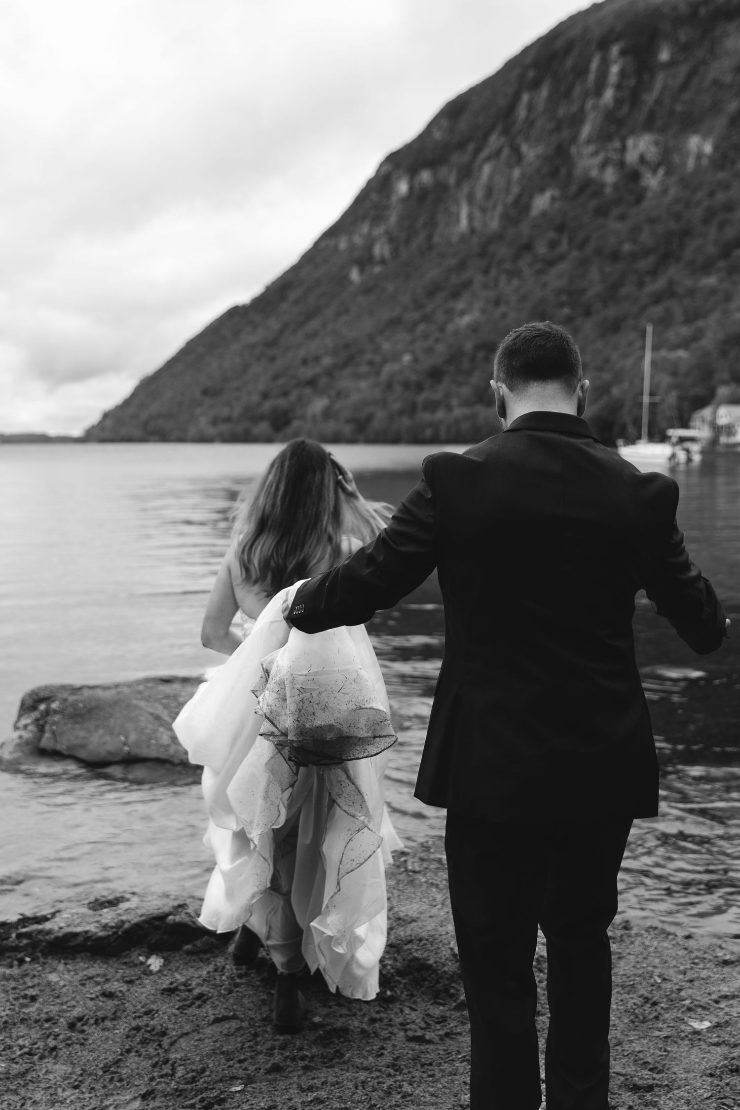 Black and white photo of couple walking along rocky lakeshore with mountain backdrop.