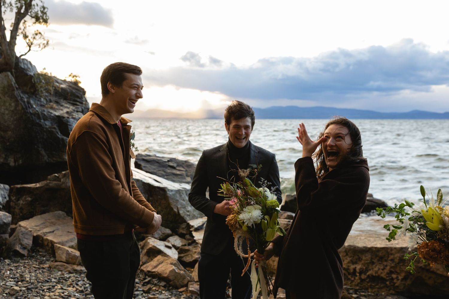 Three people gather on a rocky beach coastline at sunset holding bouquets of flowers by the ocean waves.