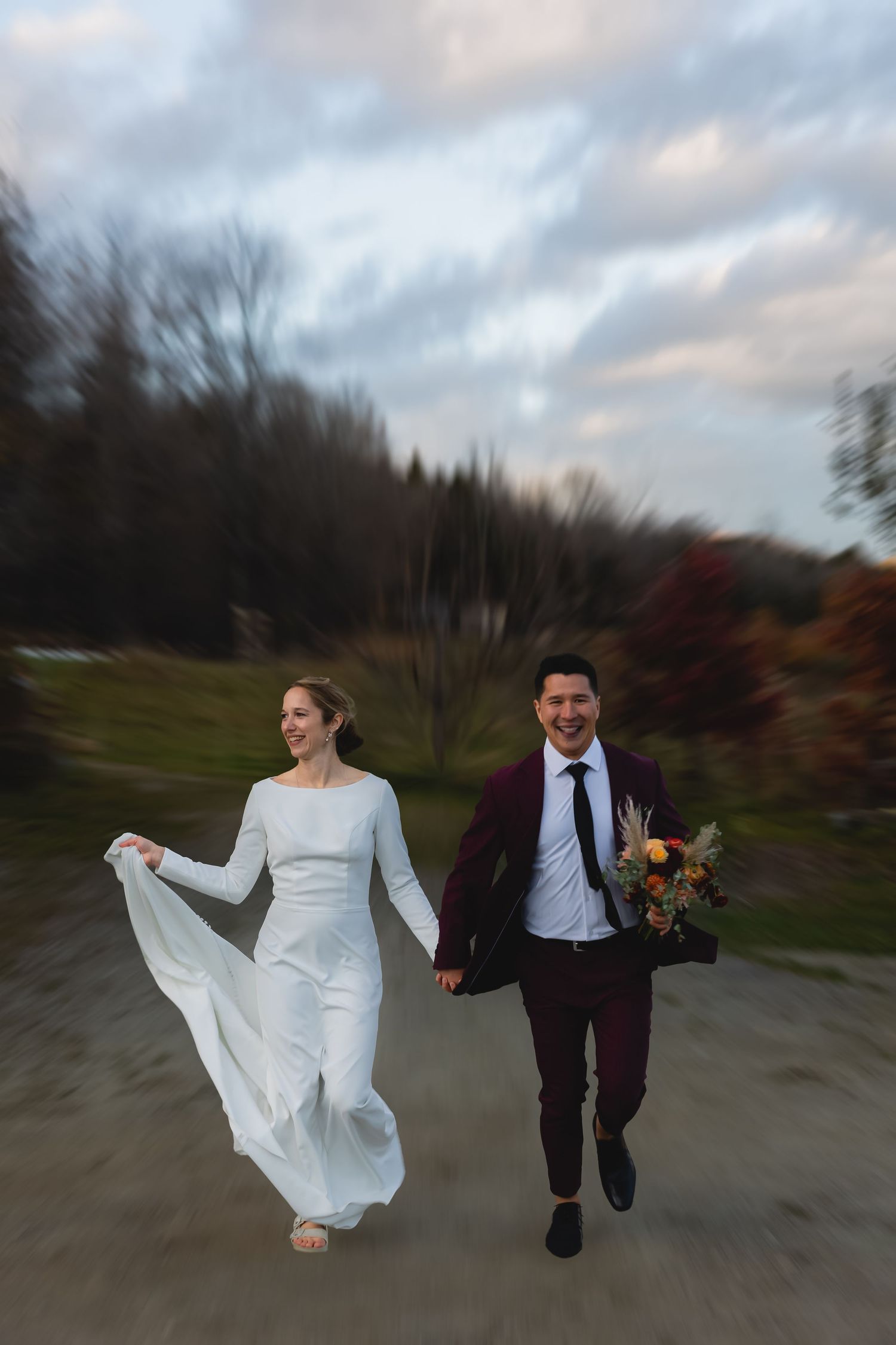 A couple in wedding attire runs joyfully down a path surrounded by autumn trees during their wedding celebration.