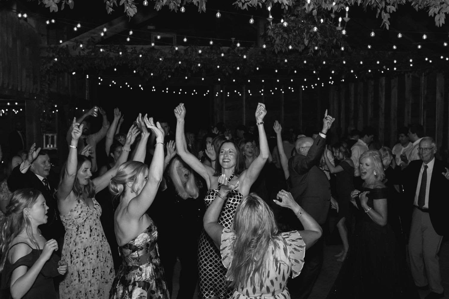 Black and white photo of a dance party under string lights with a crowd dancing with raised arms.