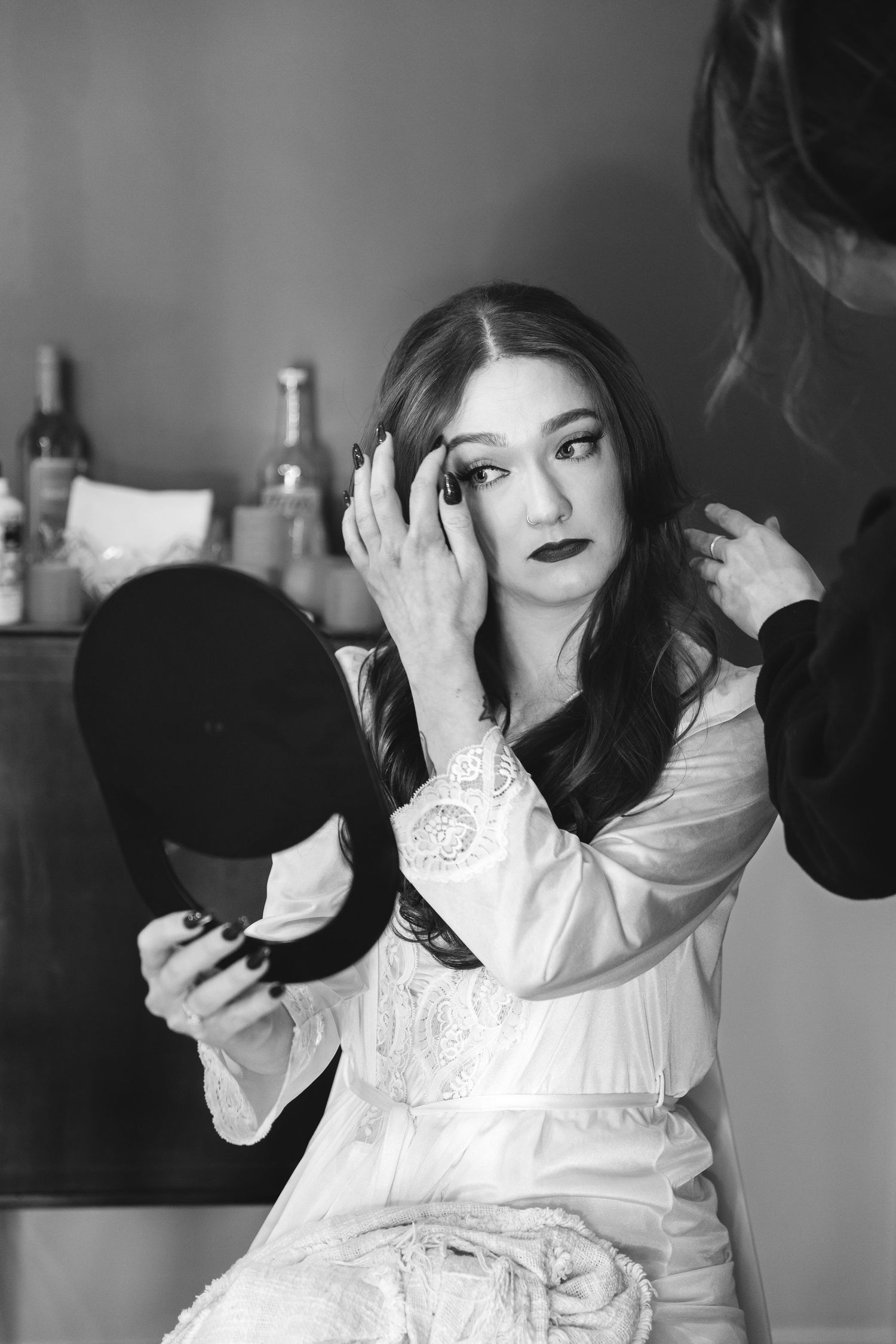 Black and white photo of someone getting ready at a vanity table wearing a white lace robe while holding a makeup mirror.
