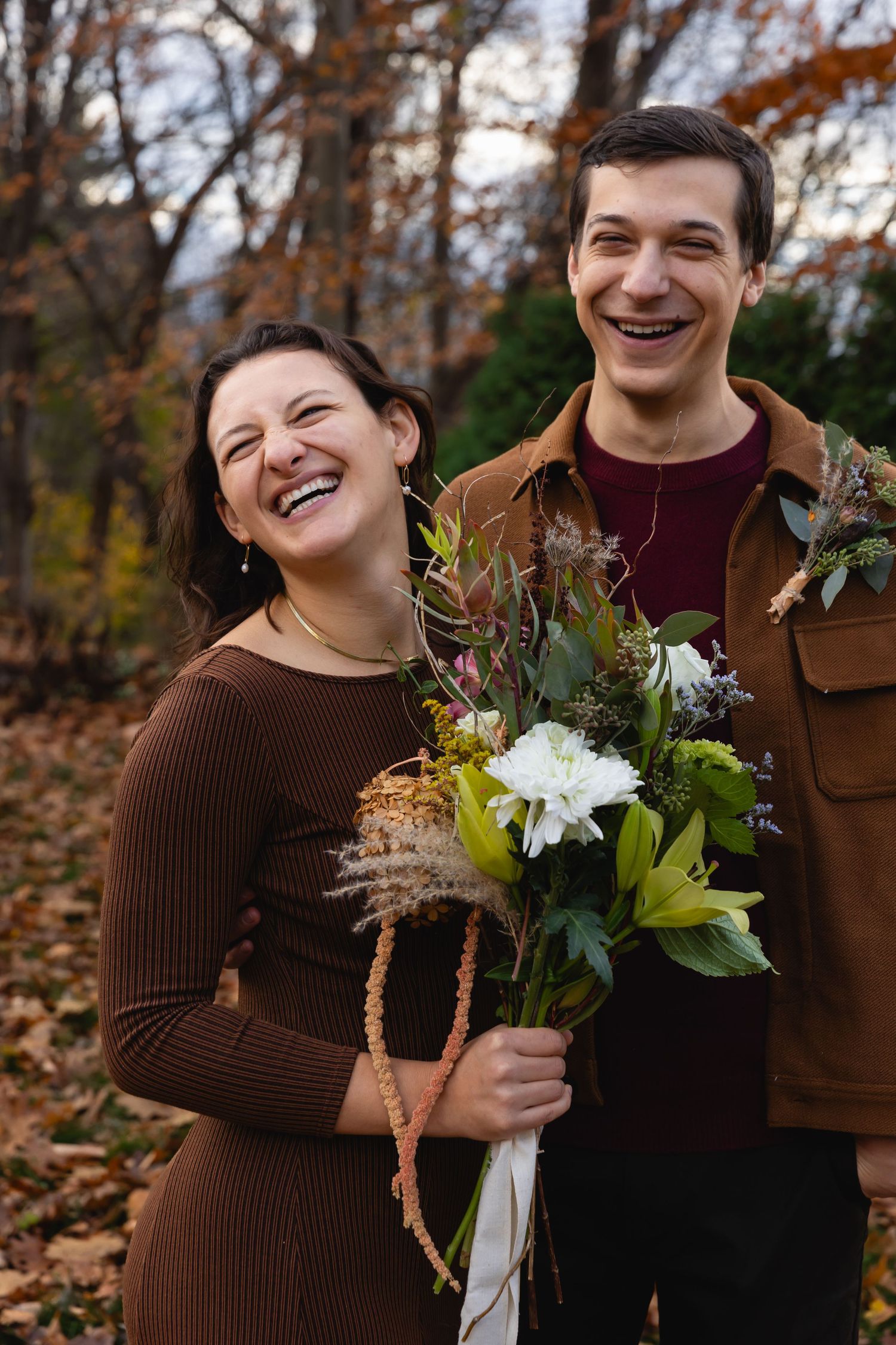 A joyful couple laughs together in autumn forest scenery while holding a bouquet with white flowers and greenery.