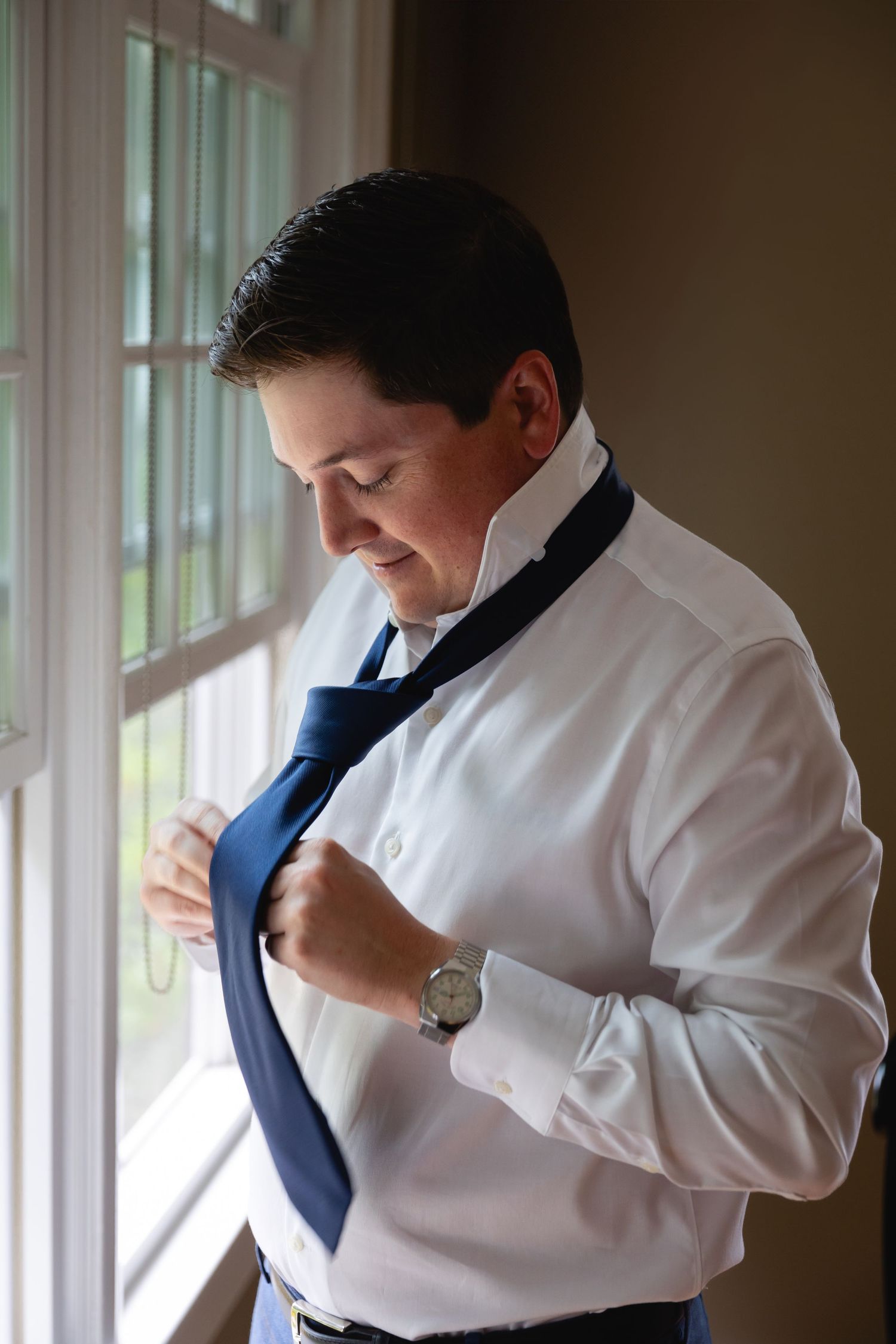 Someone adjusts their navy blue necktie while wearing a crisp white dress shirt by a window in natural light.