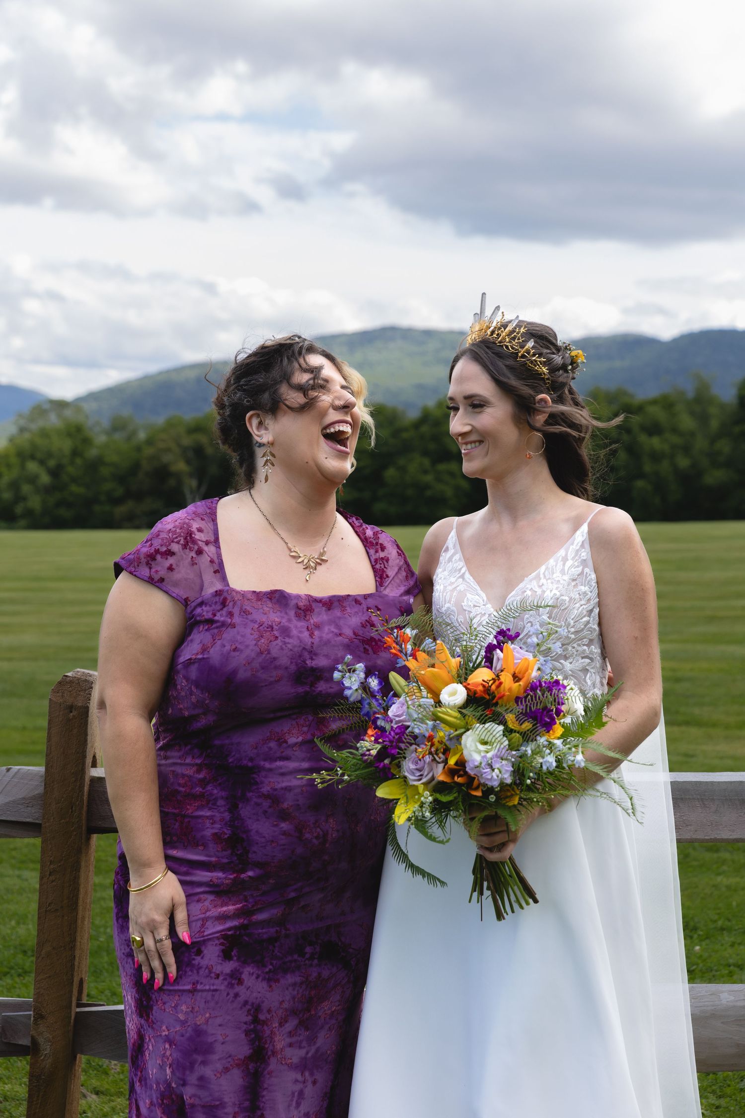 Two wedding attendees share a joyful moment against mountain backdrop with colorful bouquet.