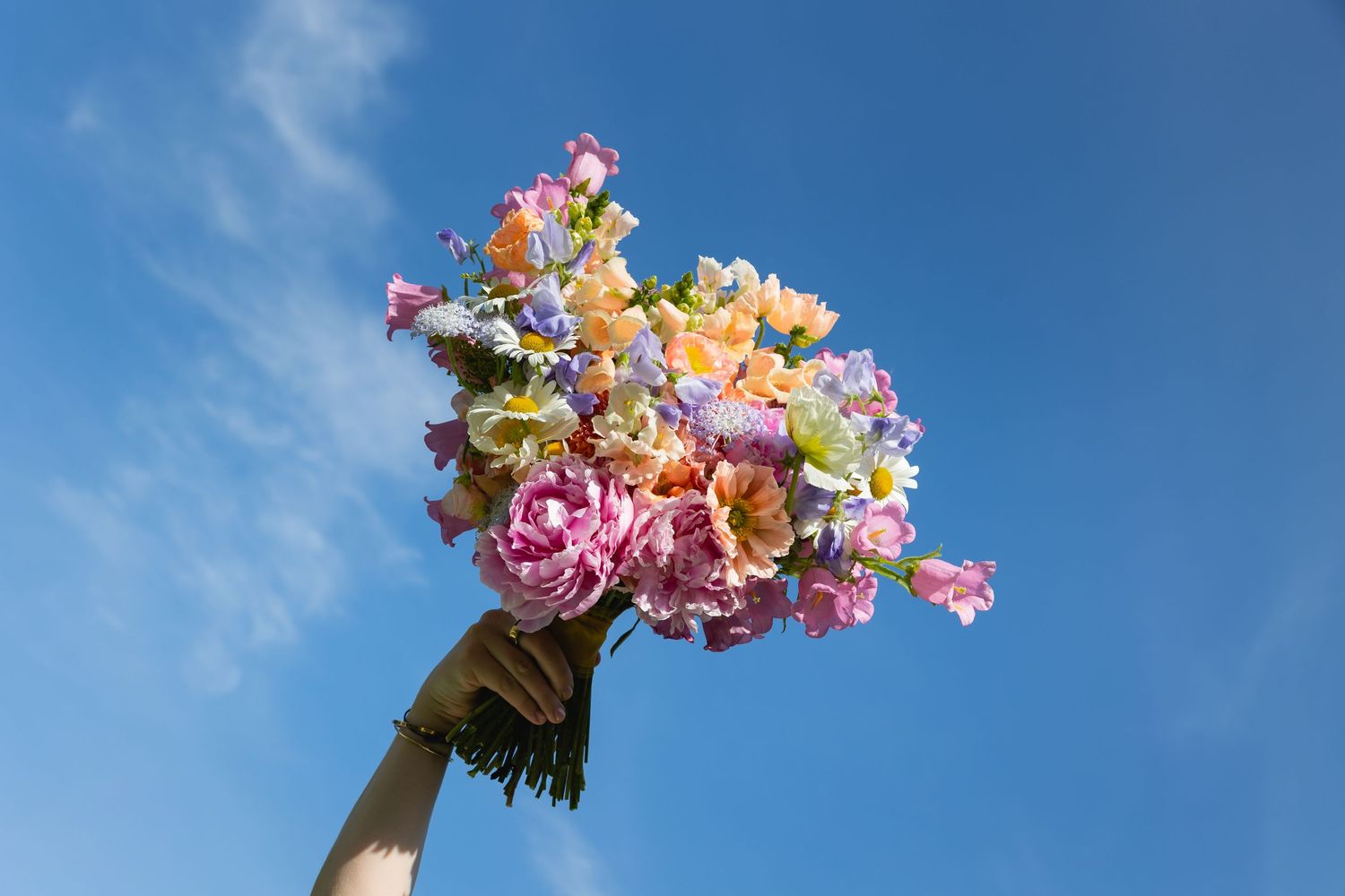 Hand holding up a vibrant bouquet of pink and orange flowers against a bright blue summer sky.