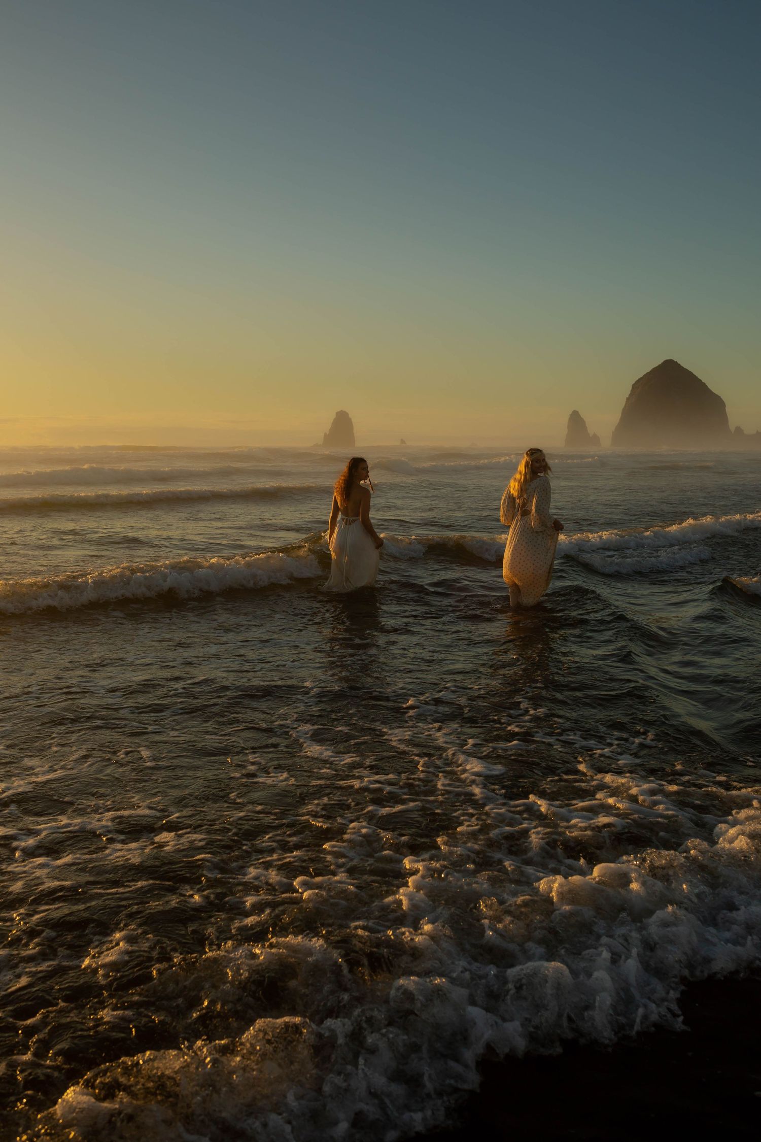 Two brides in white gowns wade into ocean waves at golden sunset near Haystack Rock at Cannon Beach Oregon.