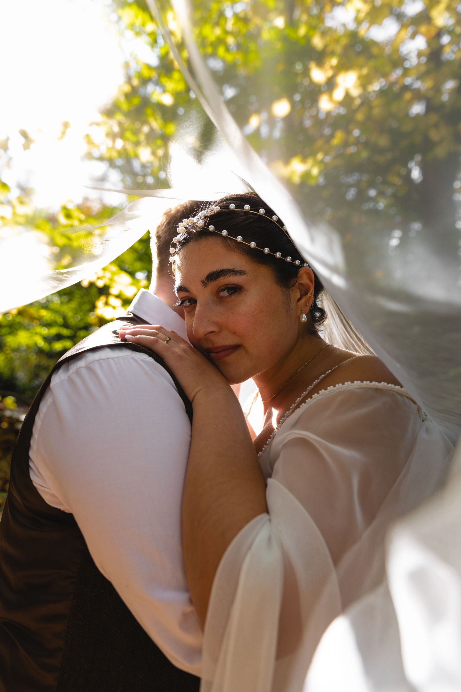 Intimate wedding portrait with flowing veil and natural sunlight in forest setting.