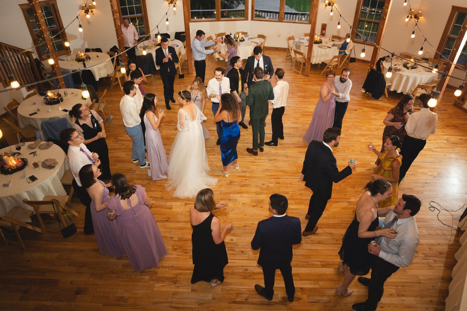 Wedding guests dancing on a wooden dance floor under string lights at an indoor reception venue.