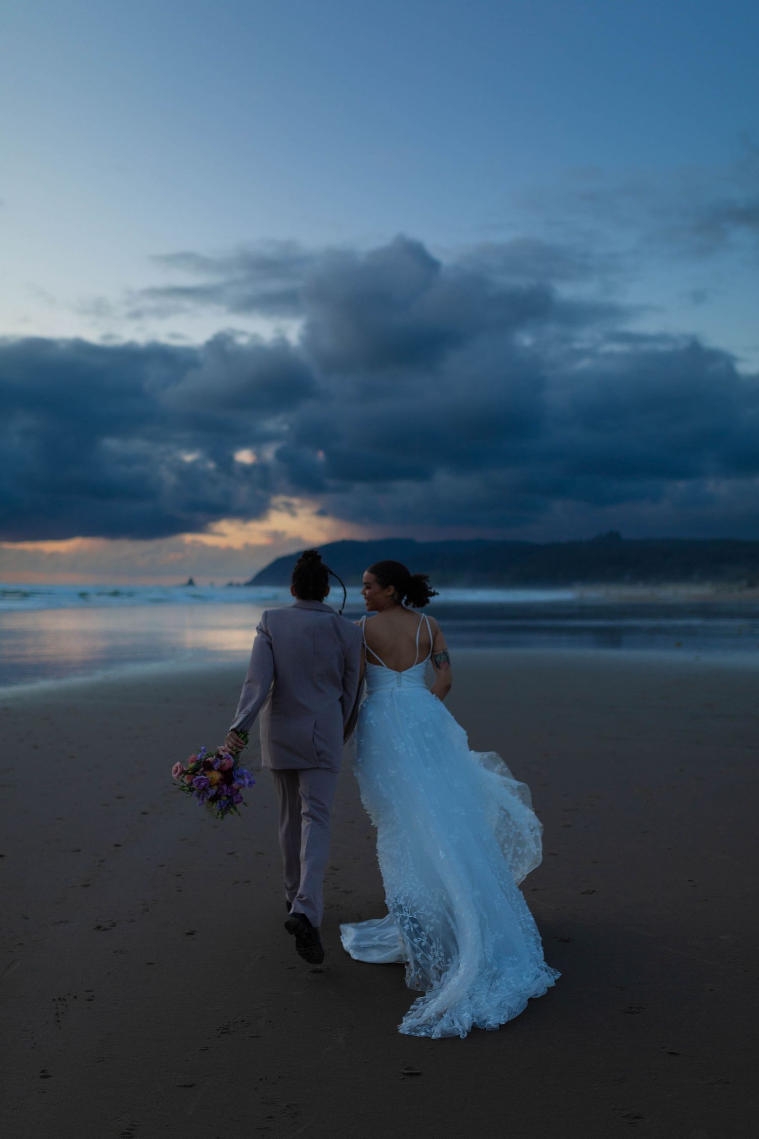 A couple walks away from camera along beach at sunset, white dress flowing in the wind.