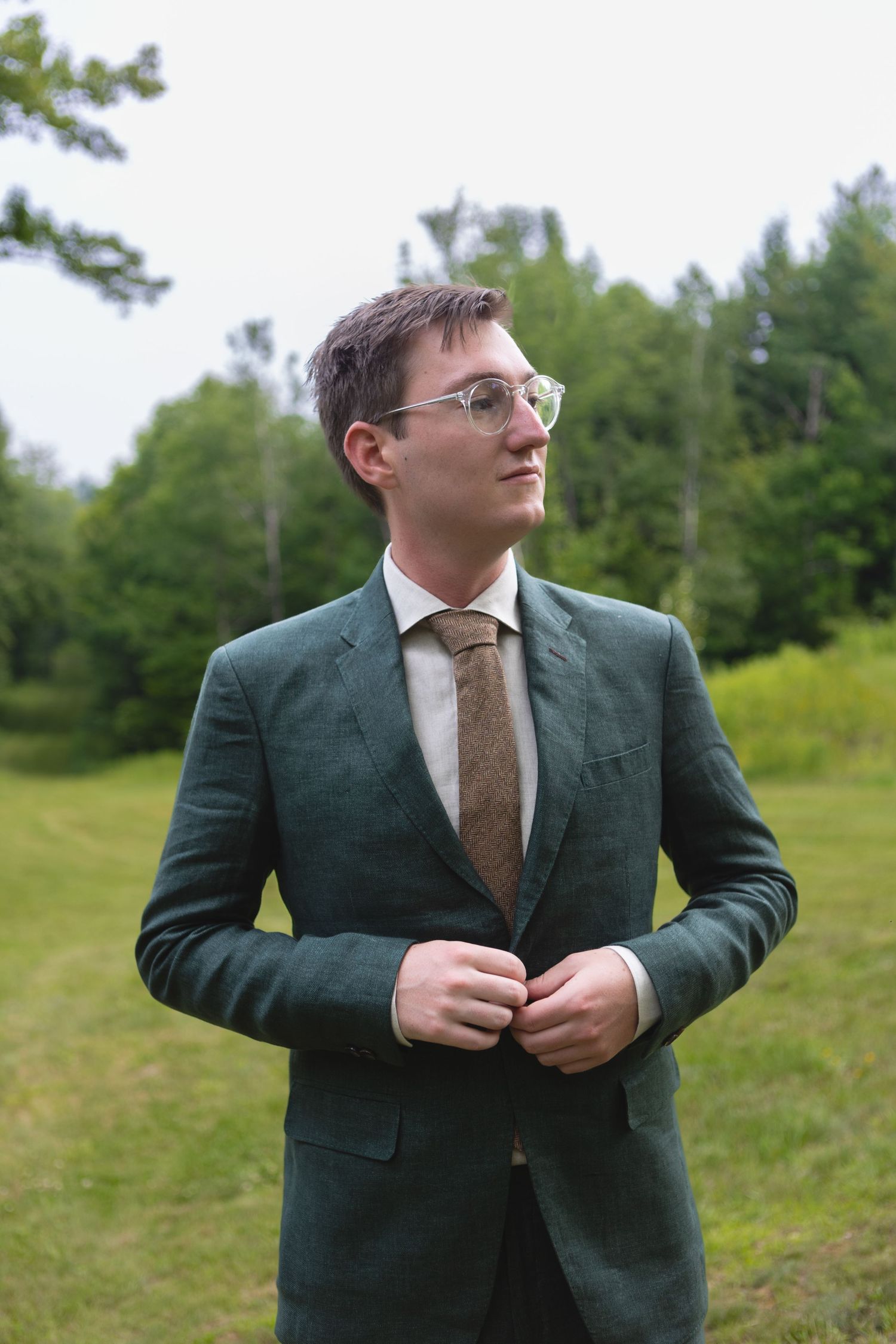 A groom in a forest green suit and glasses stands outdoors on a grassy hill.