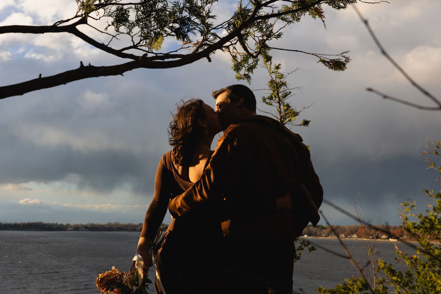 Couple sharing a romantic kiss silhouetted against a dramatic cloudy sky by the water.