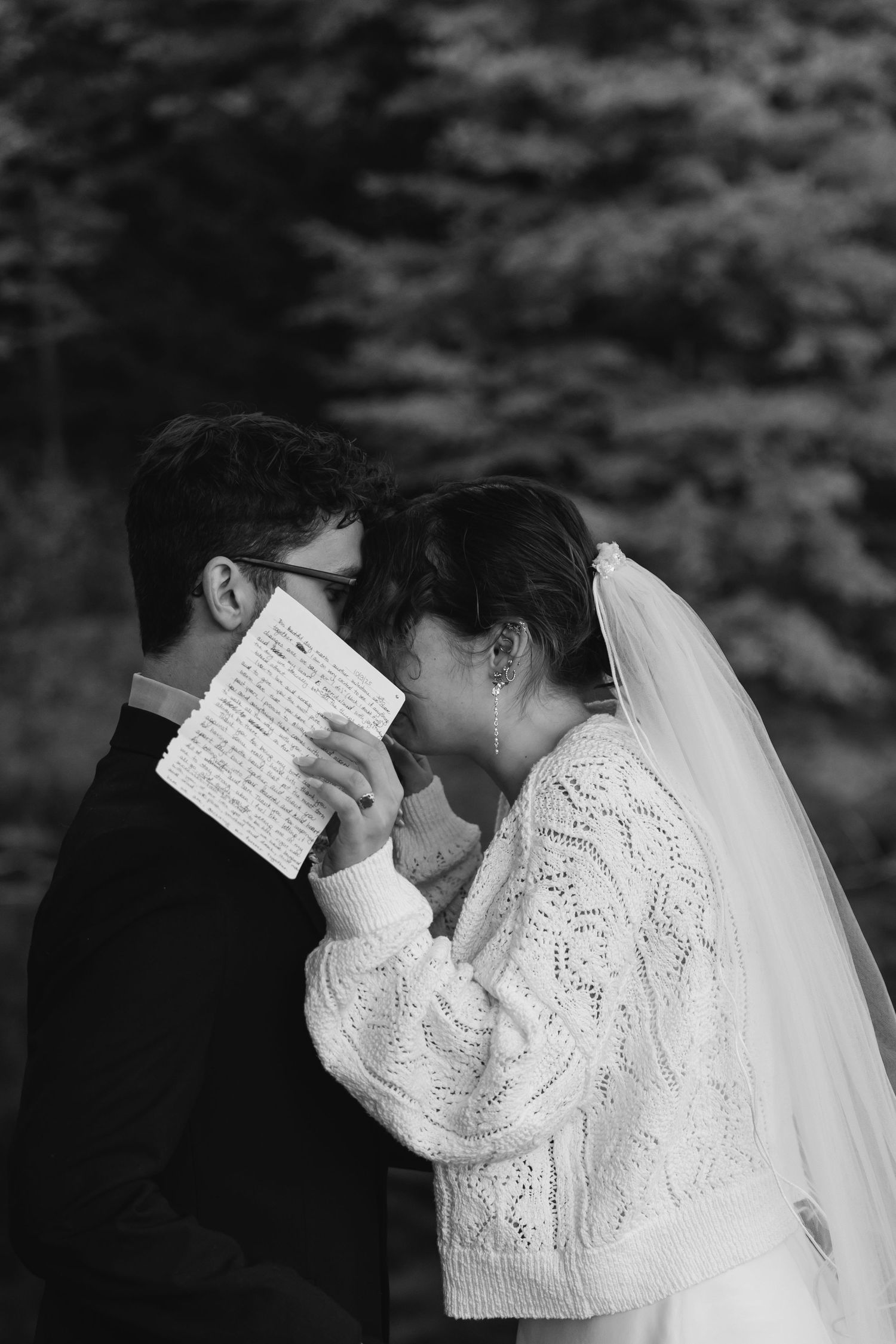 Black and white photo of a bride and groom sharing an intimate moment while reading wedding vows outdoors.