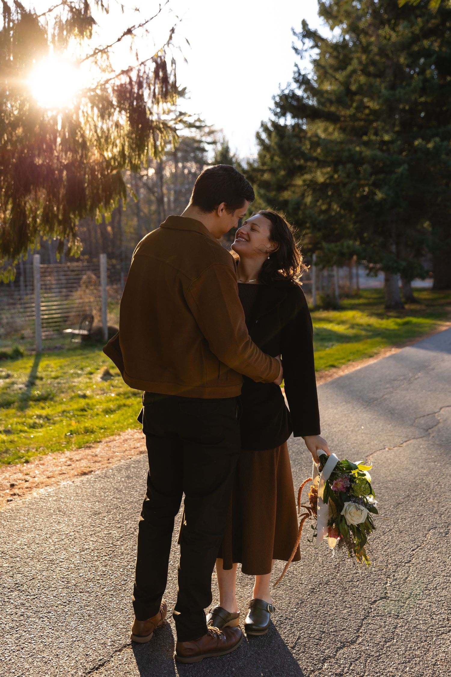 A couple shares an intimate moment on a sunlit path while holding a floral bouquet.