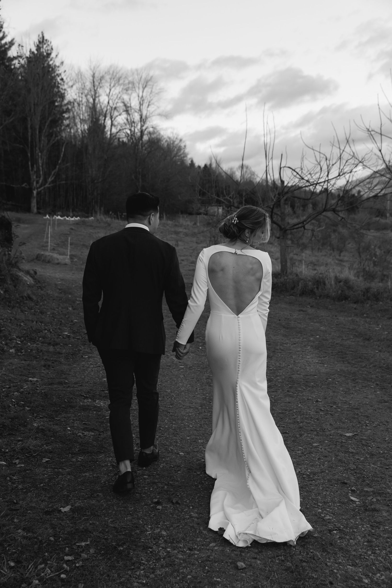 Black and white photo of a bride in backless gown and groom walking together on a rustic path with trees in background.