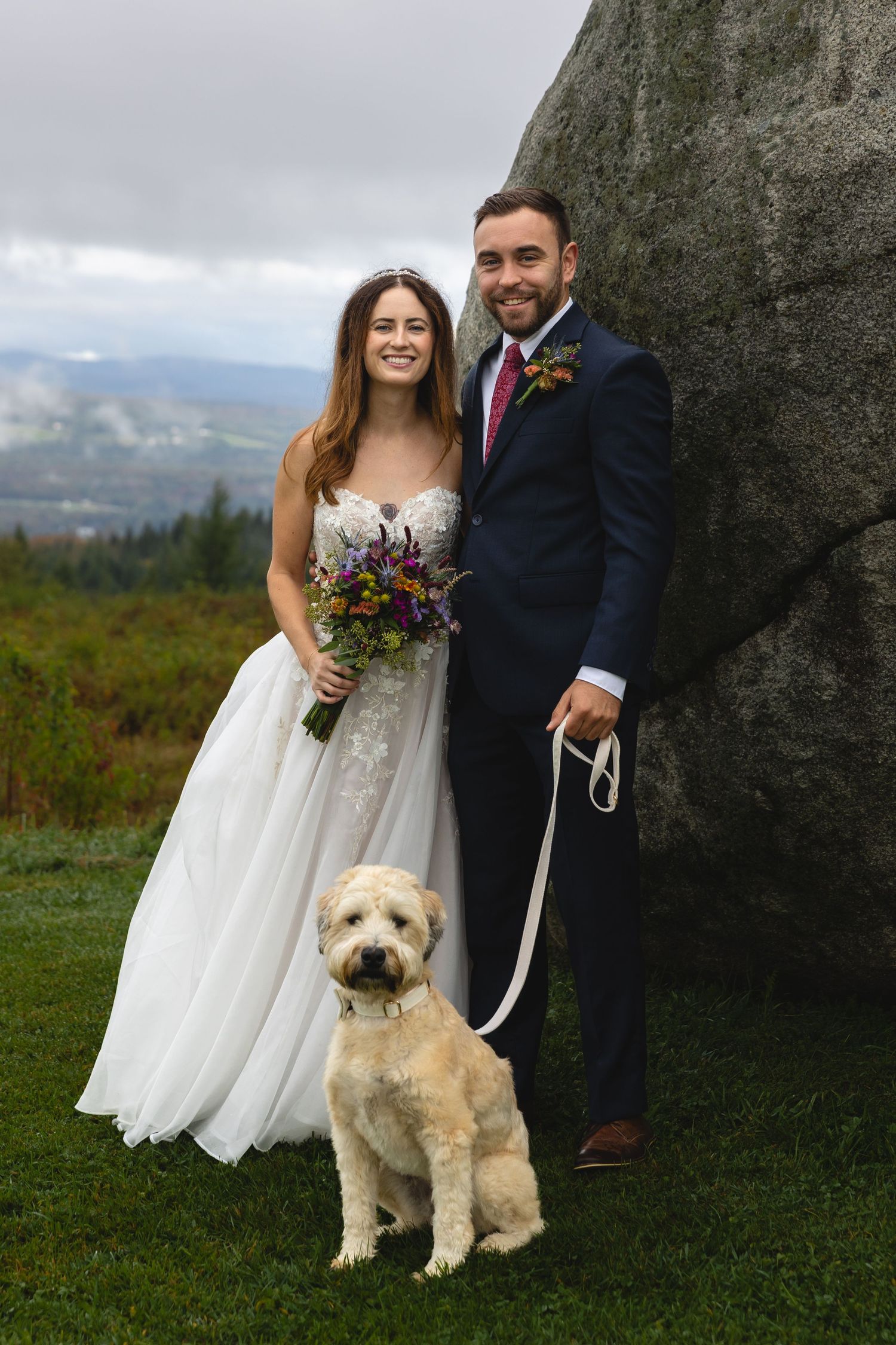 A white dog poses with a couple in formal wedding attire against a scenic mountain landscape.