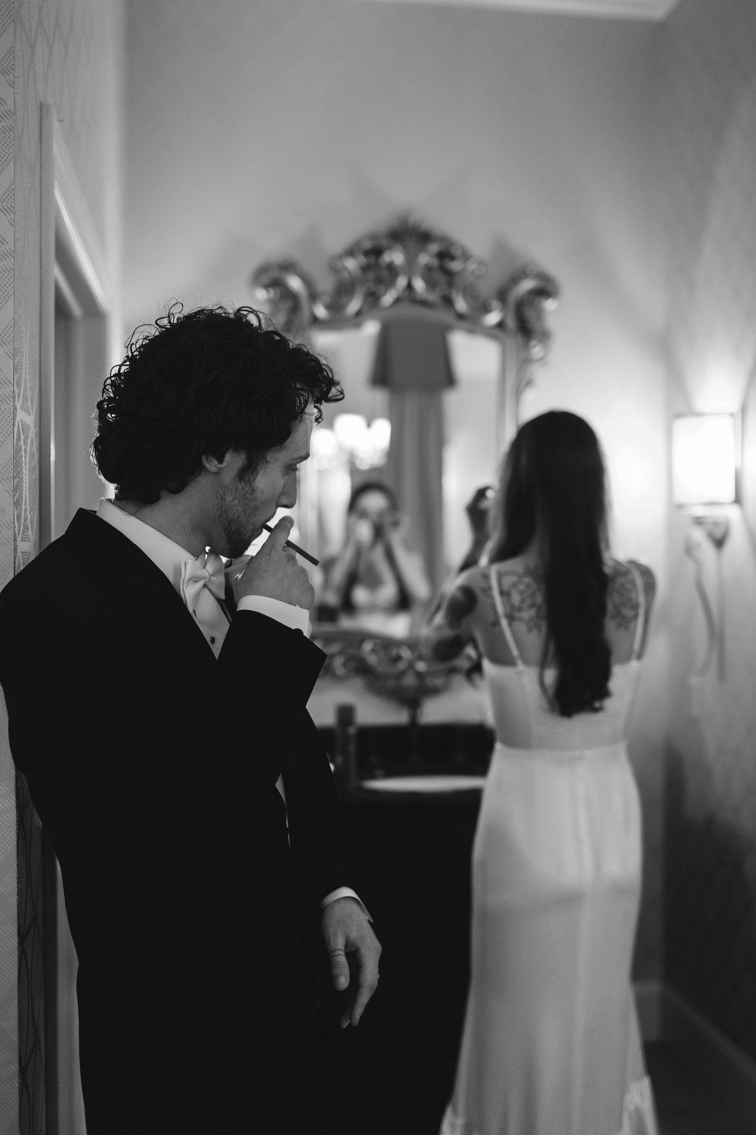 A black and white wedding photo showing a couple in formal attire standing near an ornate mirror in a hallway.