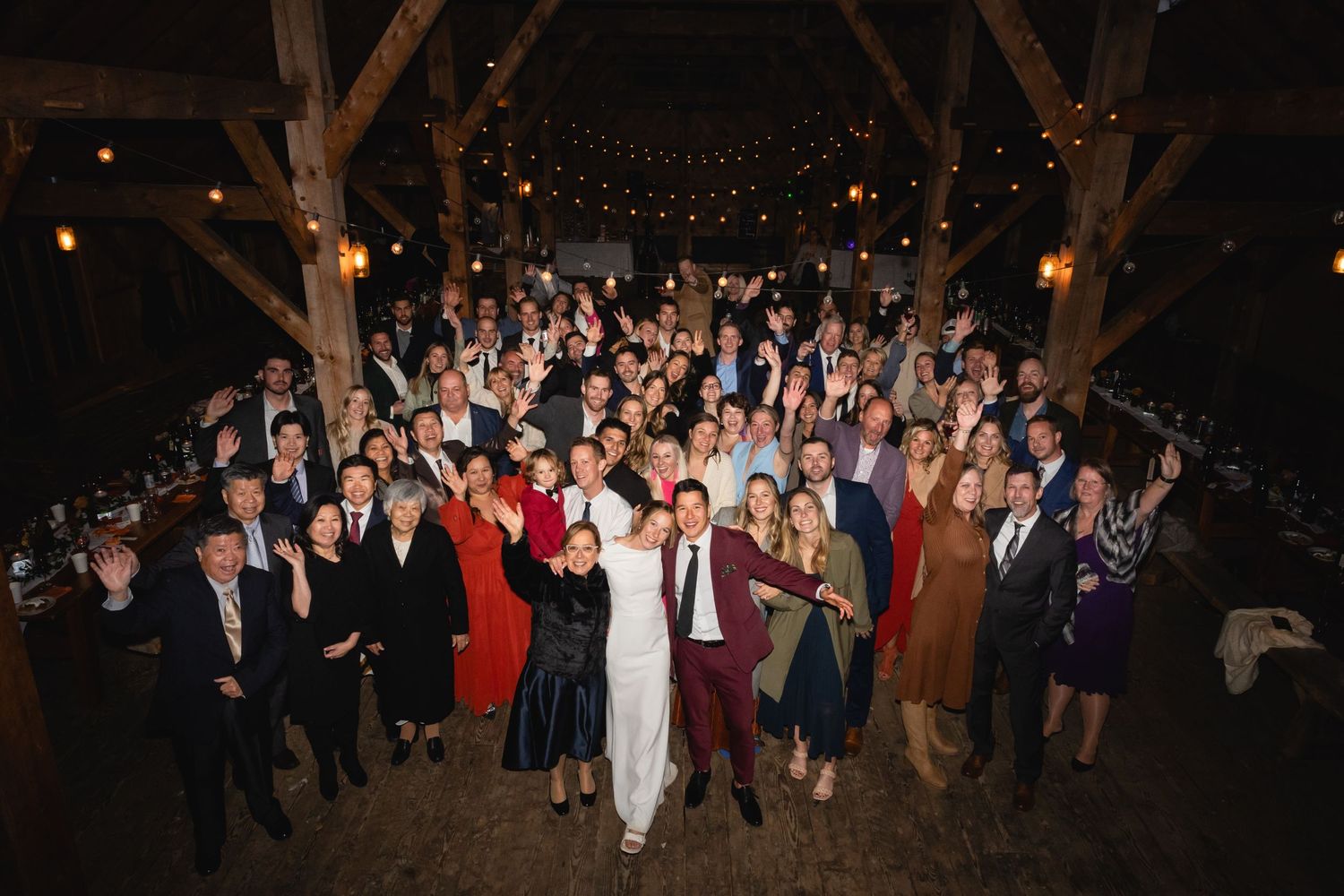 A large group of wedding guests gather under wooden beams at a dimly lit reception venue with string lights overhead.