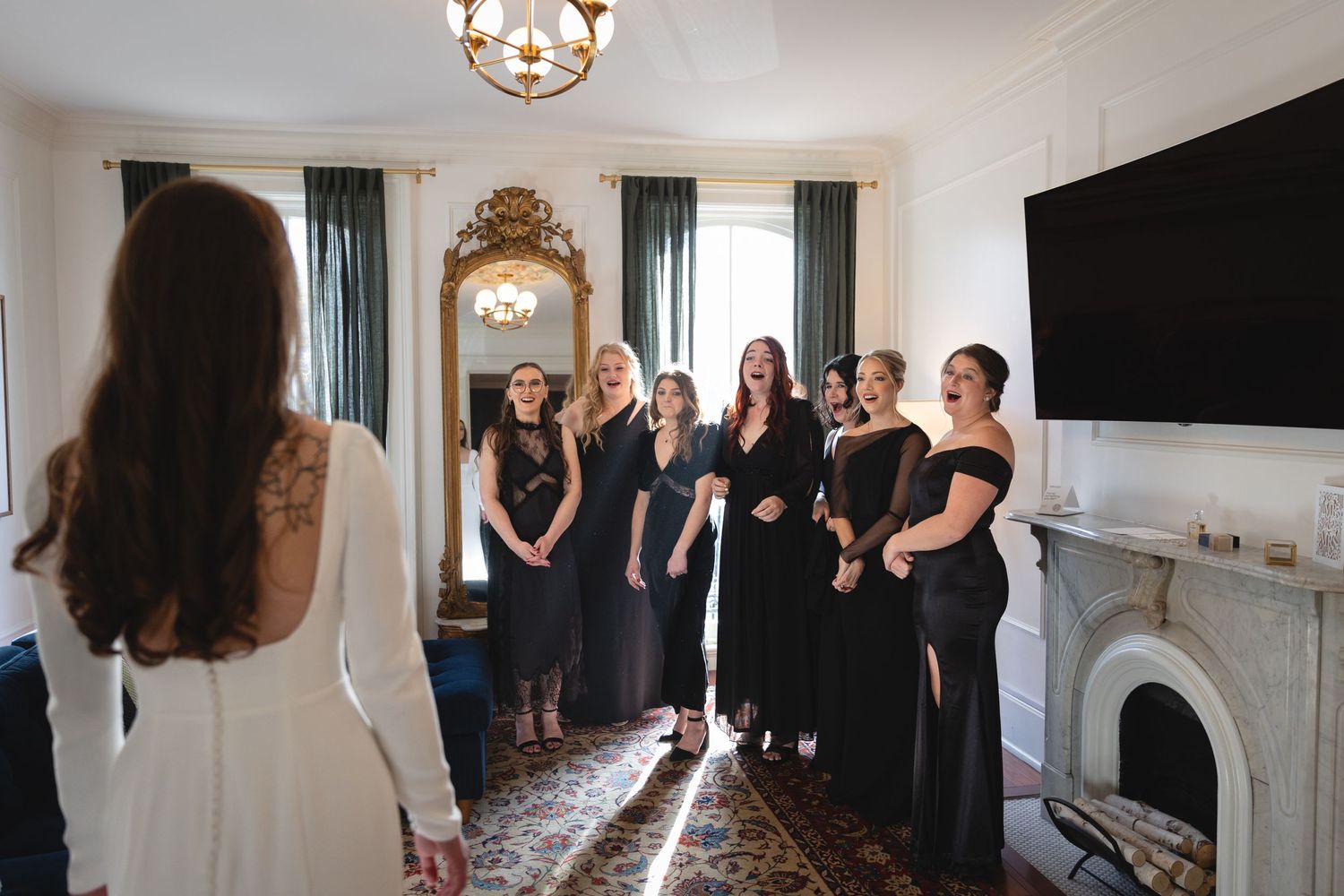 Group of women in elegant black dresses stand together in a bright room with a fireplace and large TV mounted on the wall.