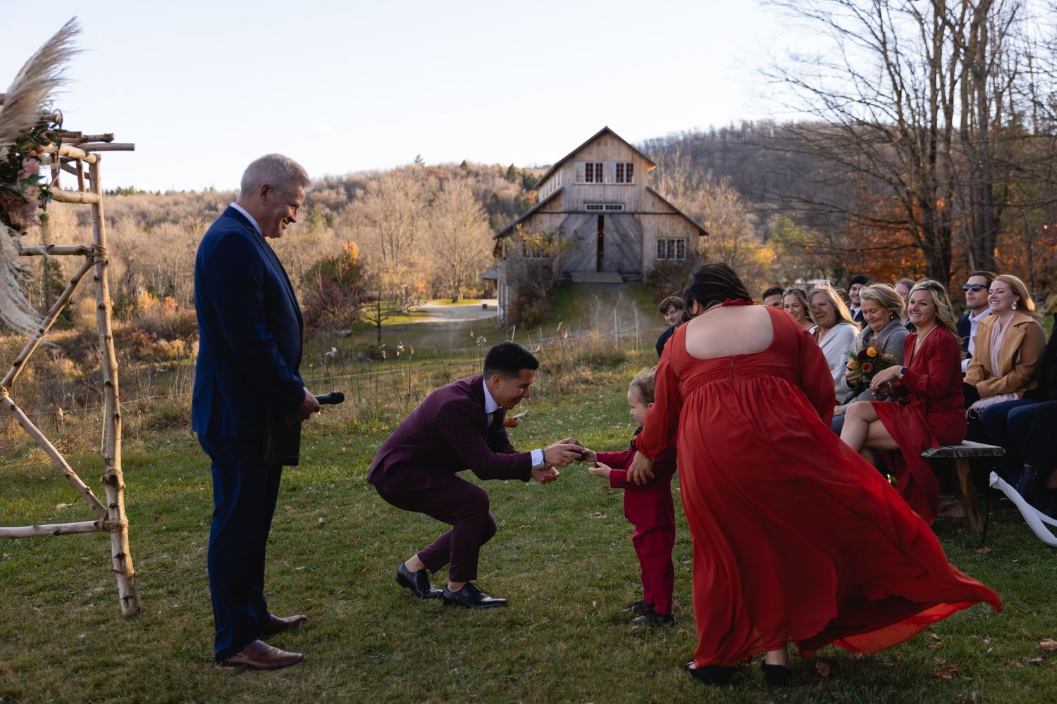 People in formal attire gather for an outdoor celebration with a rustic house and autumn hills in the background.