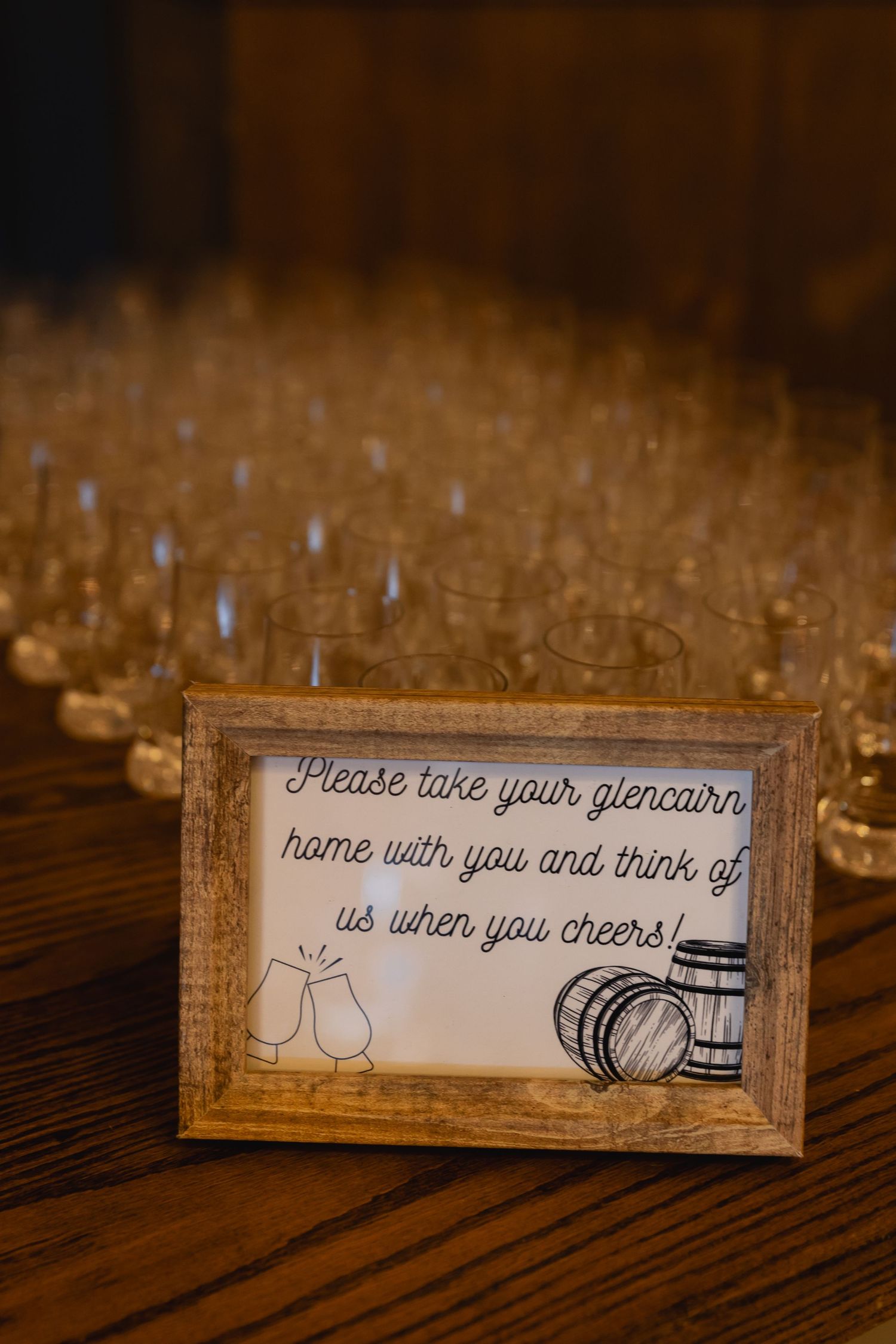 A rustic framed sign at a wedding reception invites guests to take glencairns home as favors with glasses in background.