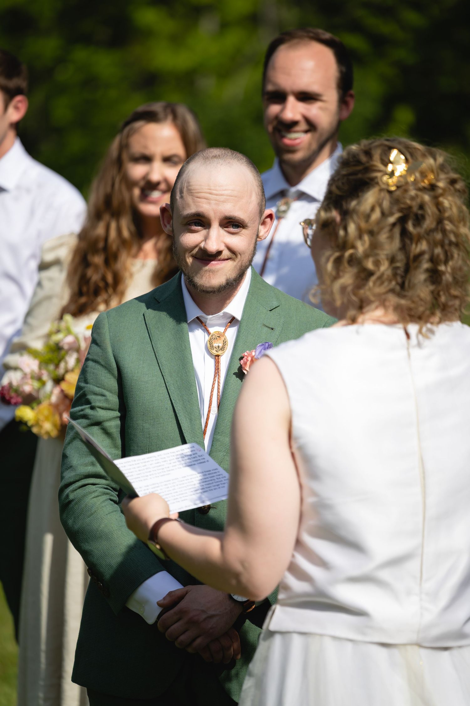 A couple exchanges vows during an outdoor wedding ceremony with guests visible in the background on a sunny day.
