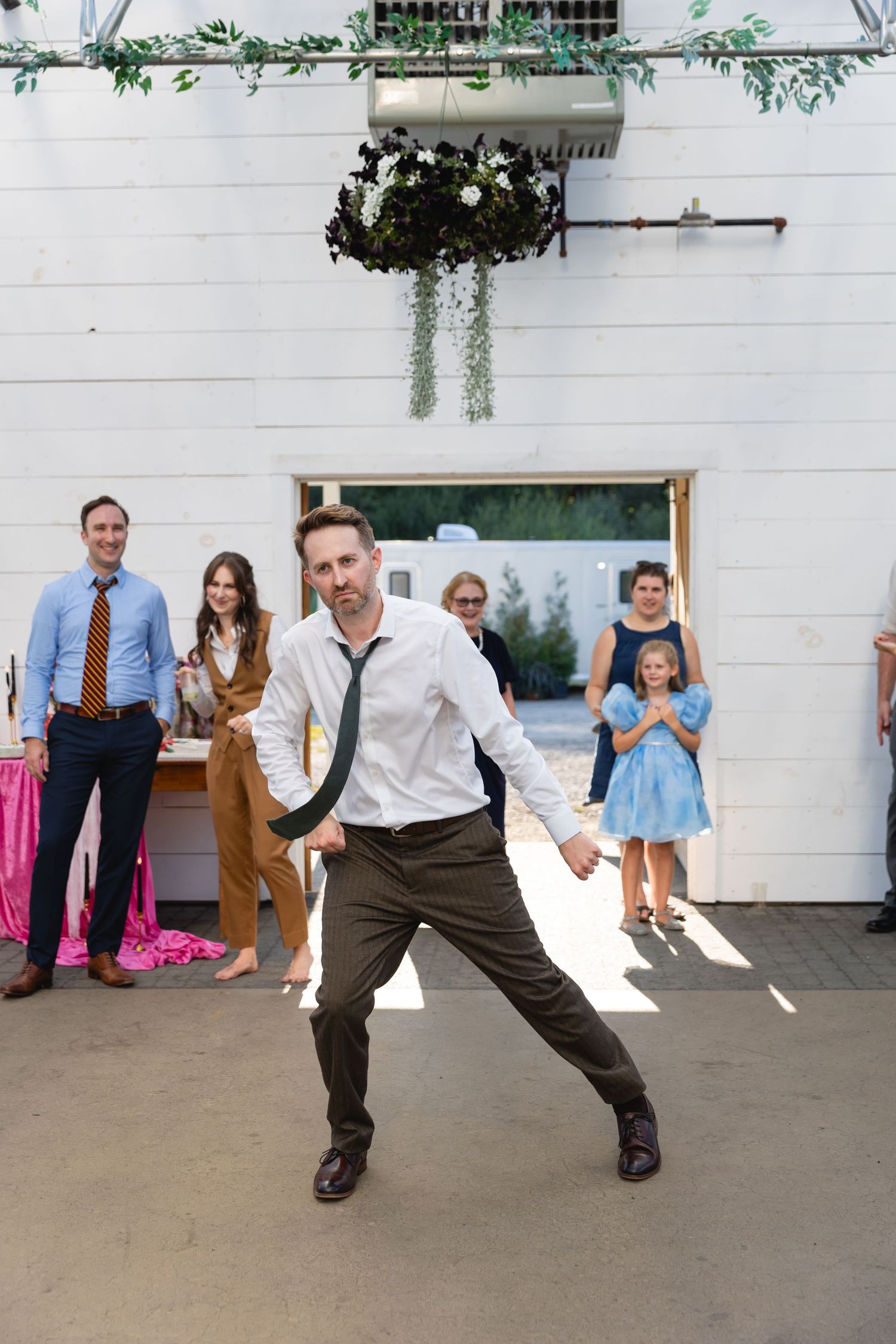 A lively dance moment captured at a casual outdoor event with greenery decorations against a white wall.