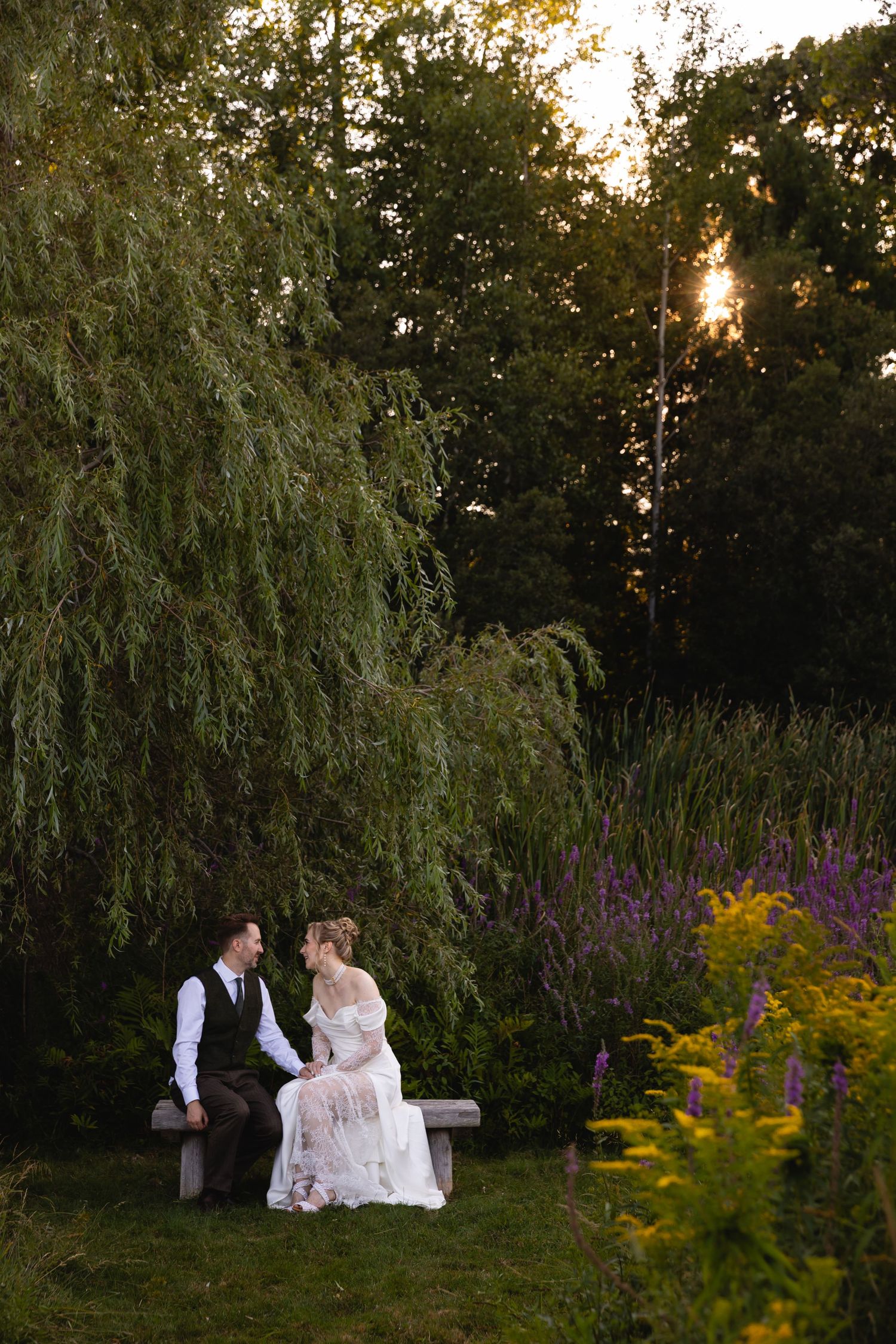 Couple sits together on bench at sunset in garden with purple wildflowers and willow trees creating romantic ambiance.