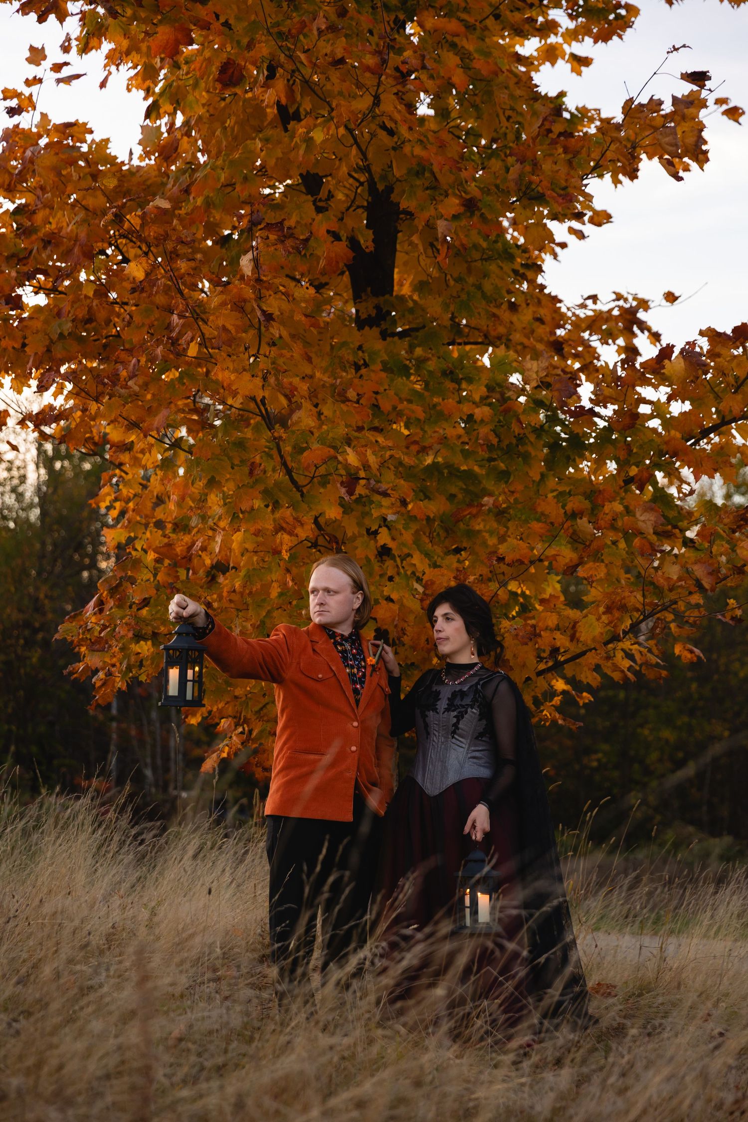 Two people stand in tall autumn grass beneath vibrant orange maple trees on an overcast day.