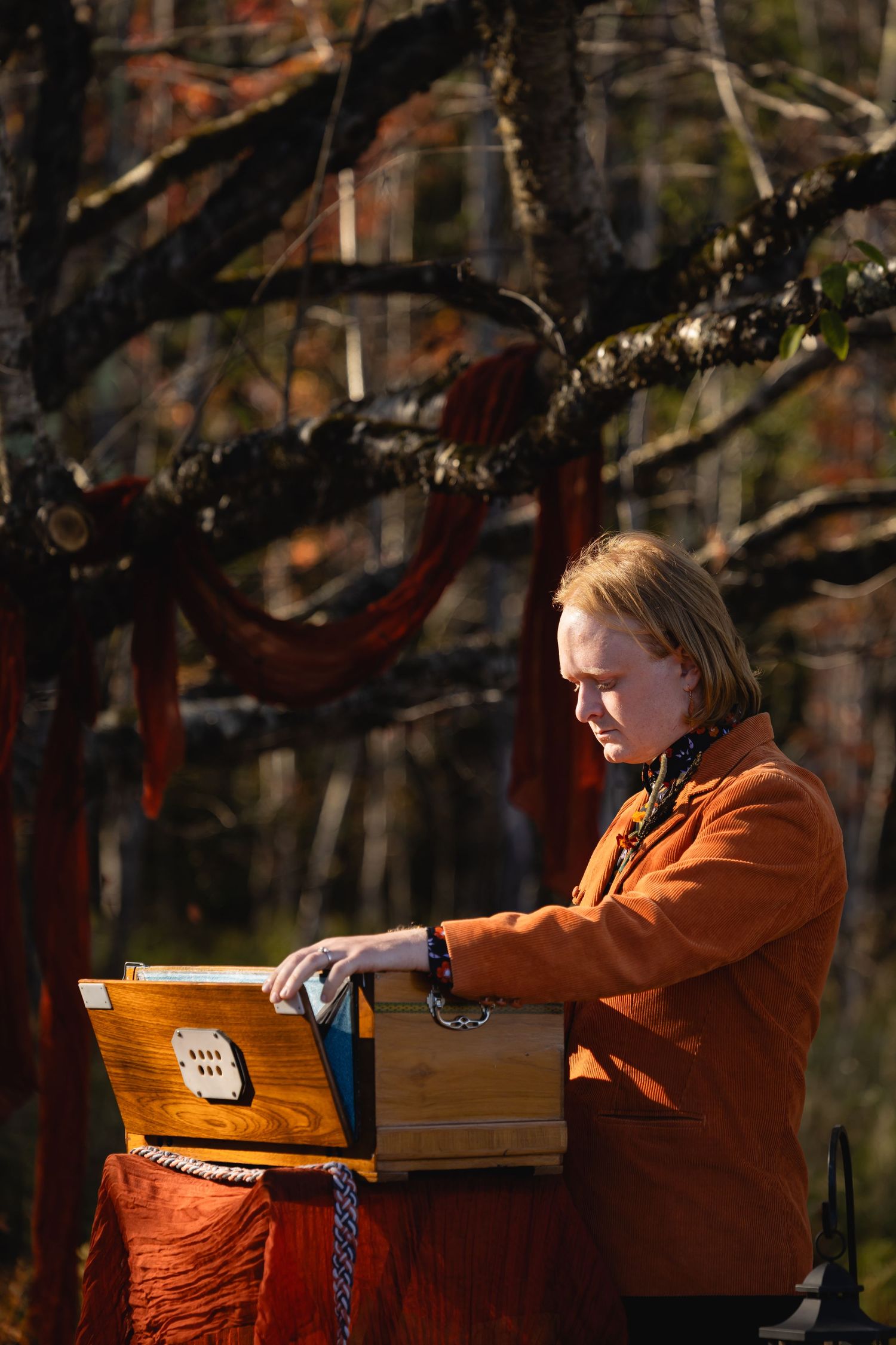 A musician in an orange jacket plays a harmonium instrument outdoors among autumn trees.