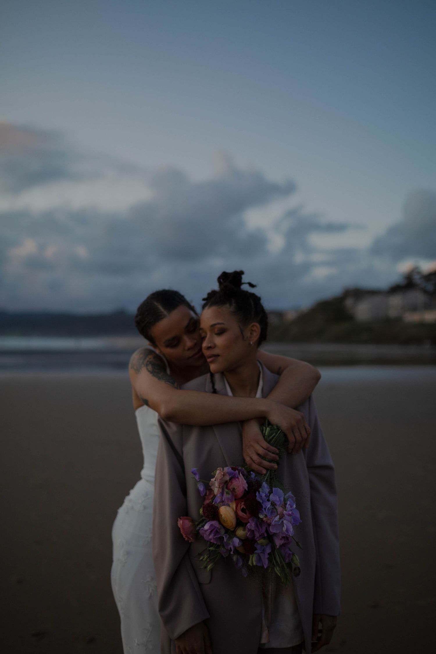 A loving embrace between newlyweds on a misty beach at twilight with a colorful bouquet.