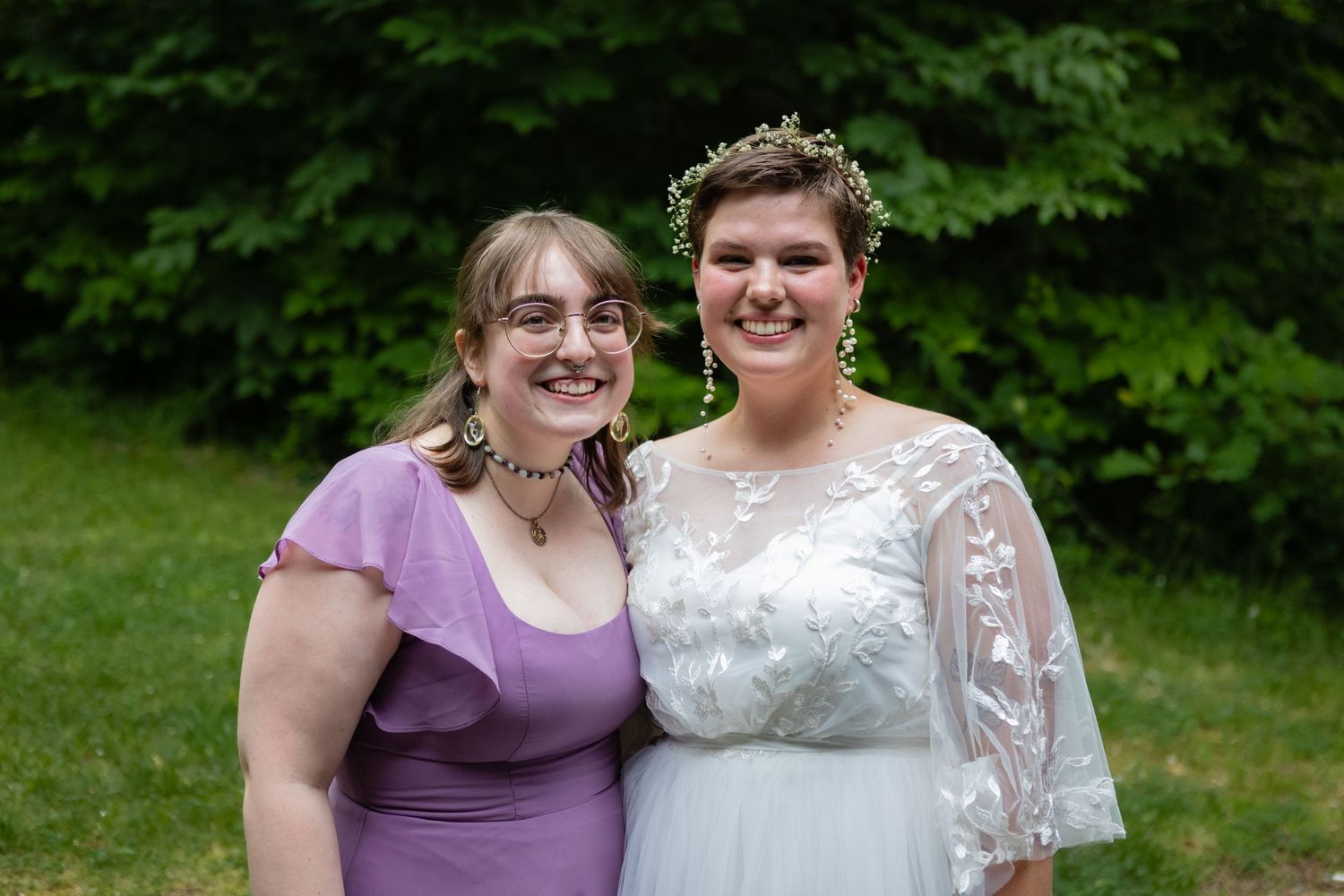 Outdoor wedding portrait with bride and guest in purple dress against stone wall and trees.