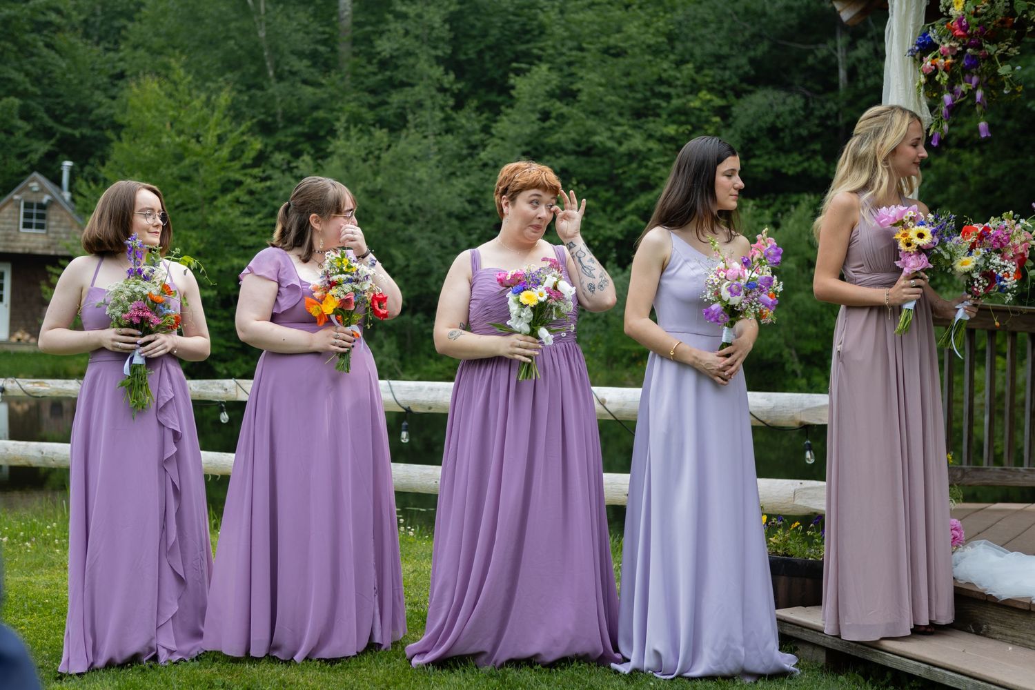 Row of bridesmaids in lavender dresses holding colorful bouquets on a wooden deck.