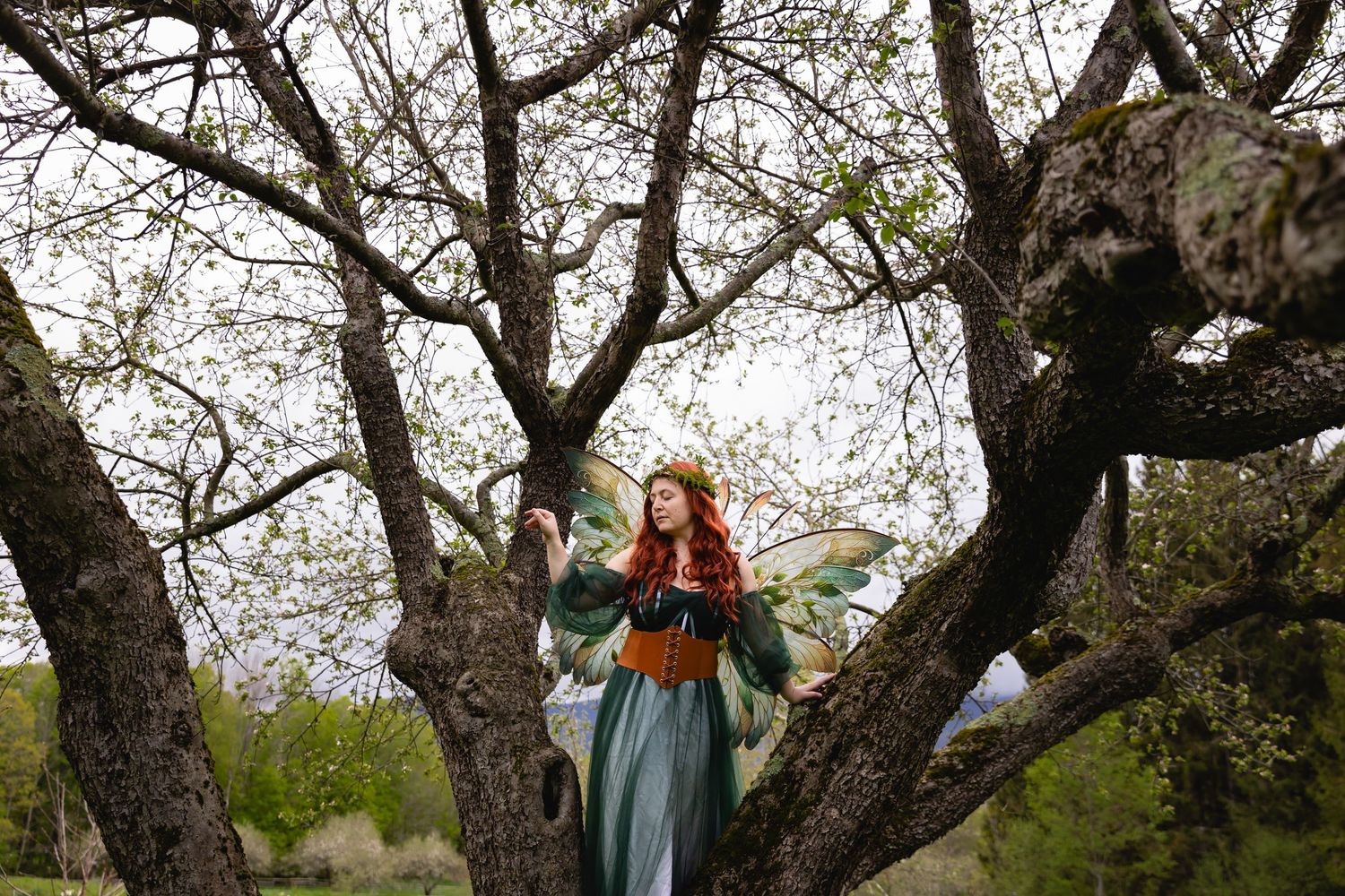 A red-haired woman in a fantasy fairy costume stands among bare branches in an orchard.
