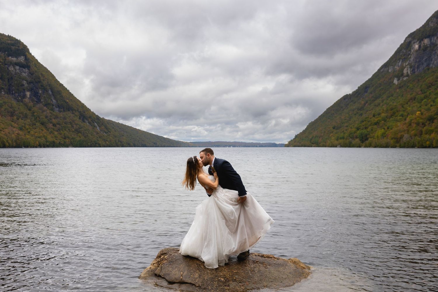 Bride and groom share a romantic dip kiss on a rock in a mountain lake under cloudy skies.