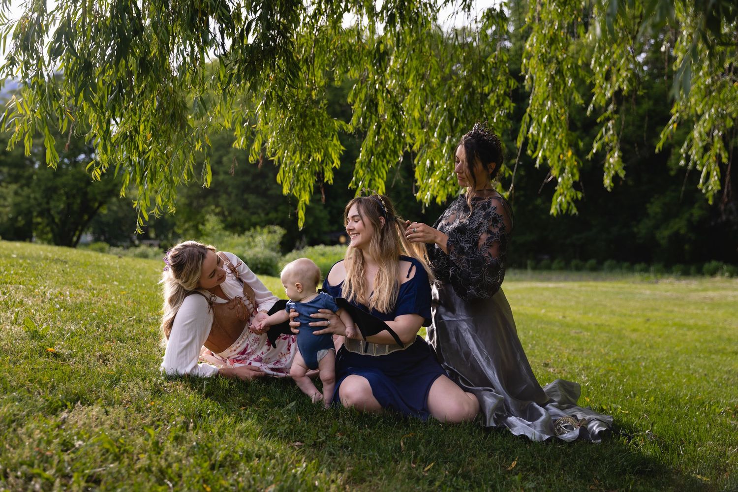 Group of women sitting under willow tree in sunny park with baby.