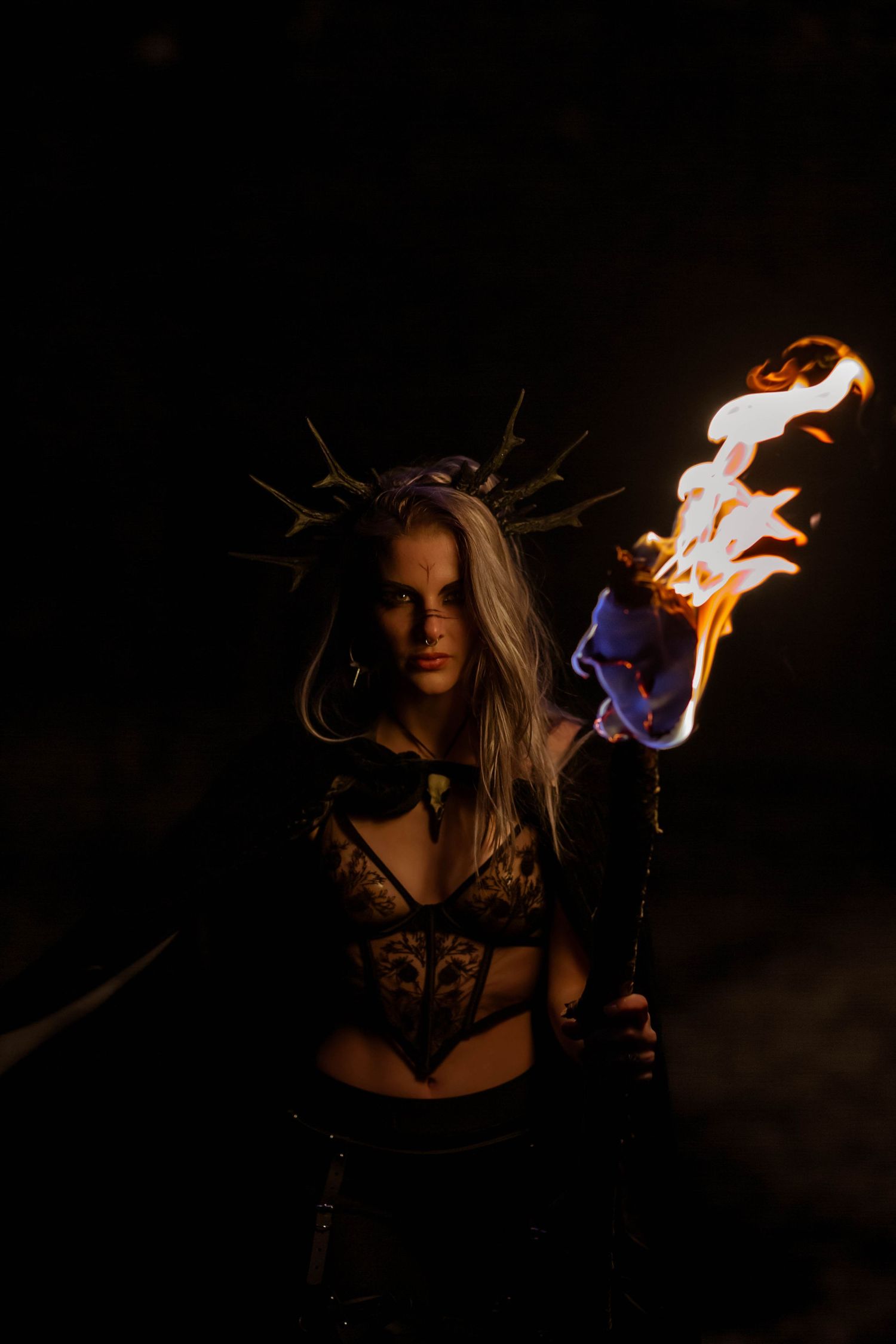 Fierce woman in dark corset costume holds flaming torch against black background.
