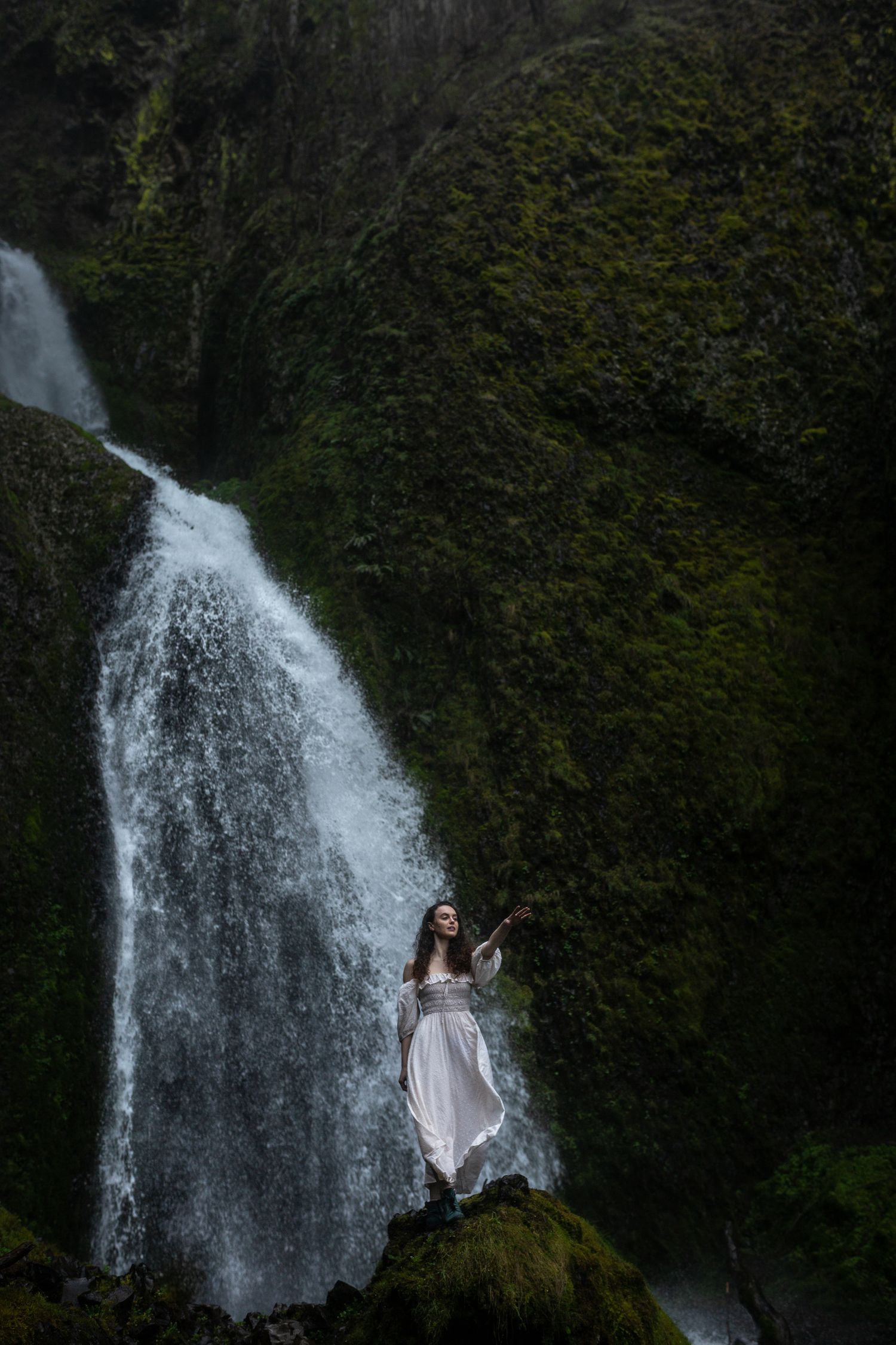 Woman in white dress standing on mossy rocks beside a tall cascading waterfall.