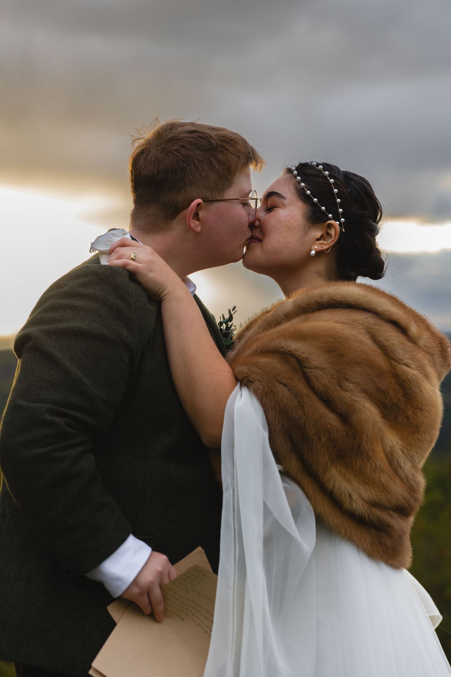 Newlyweds share an intimate kiss outdoors with dramatic cloudy sky and mountains in the background.