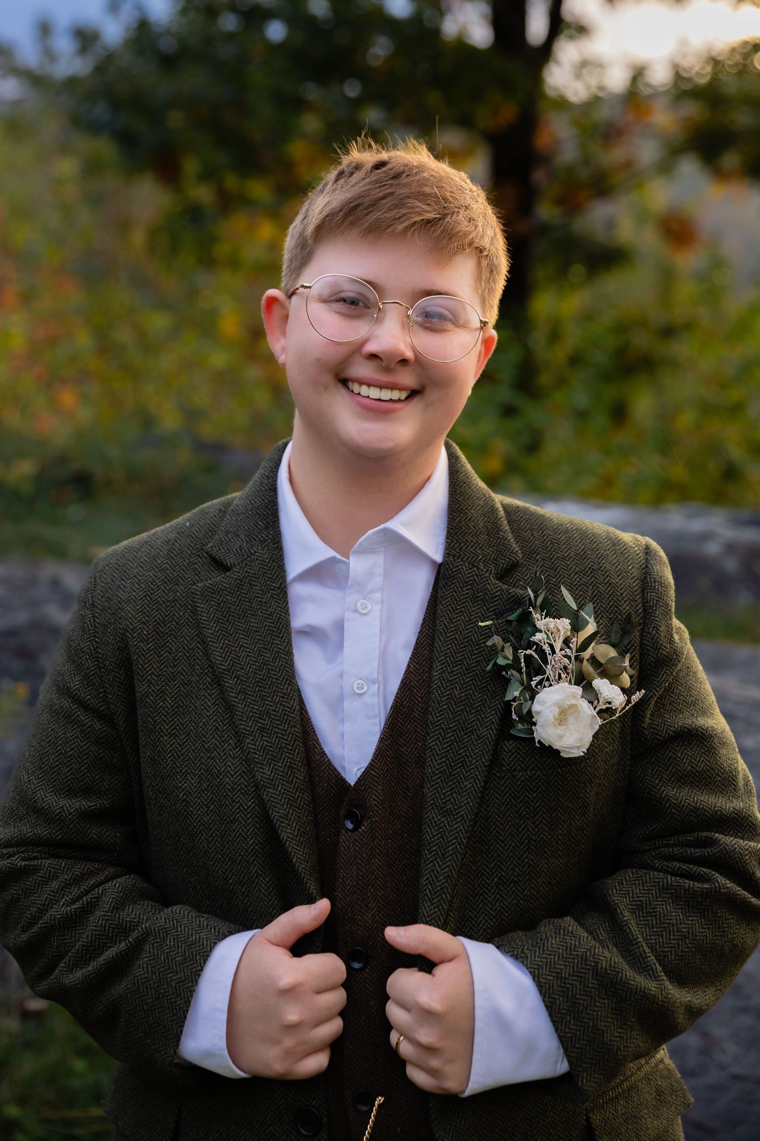 Wedding portrait of someone in a dark suit with boutonniere smiling against an autumn backdrop.