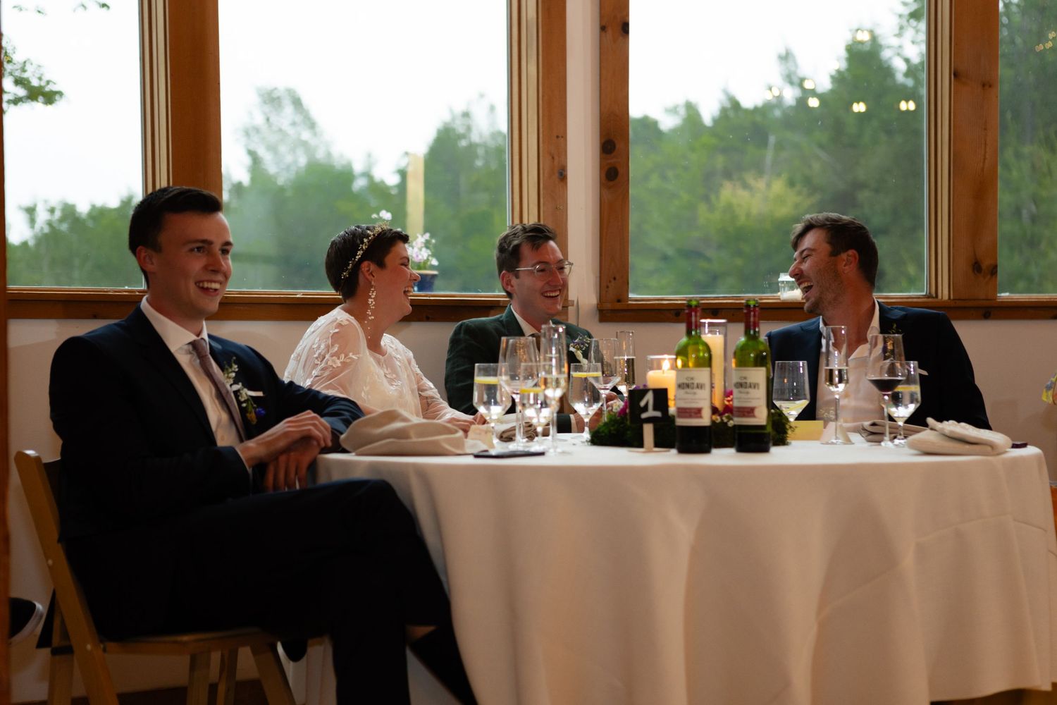 Wedding guests enjoy conversation at an elegant table setting by windows overlooking trees.