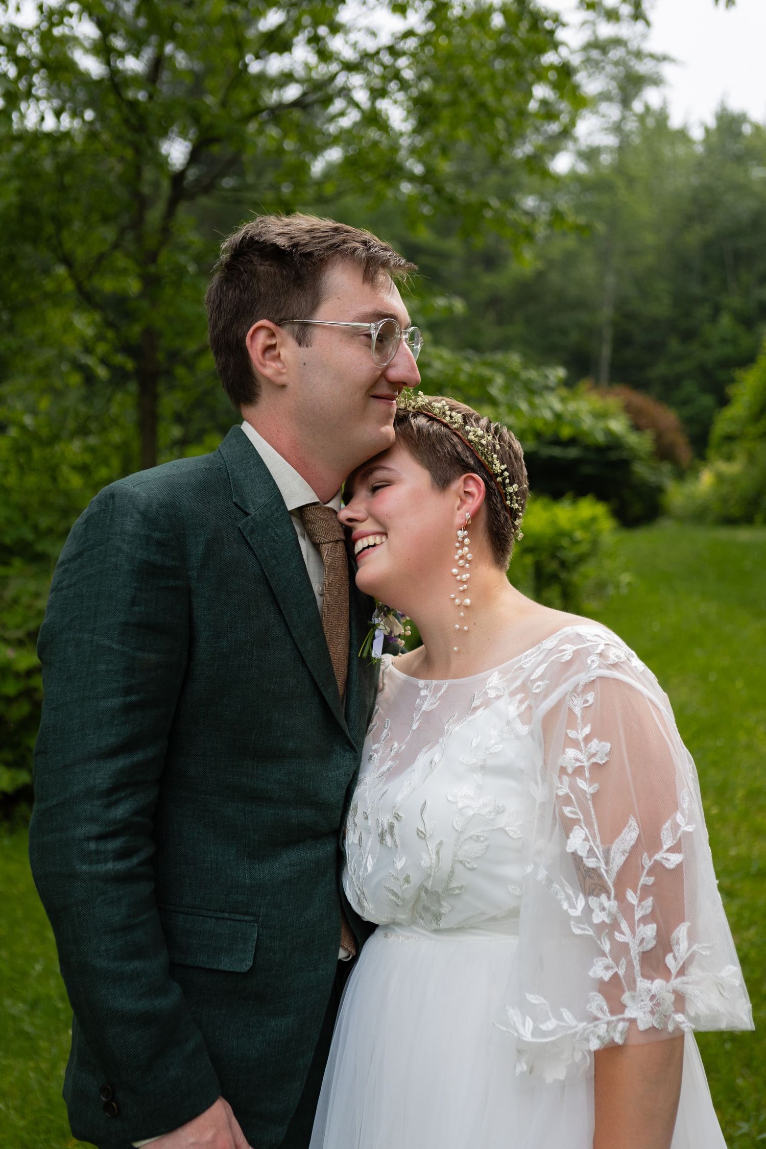 Sweet wedding moment captured in garden with bride in lace sleeves and groom in green suit.
