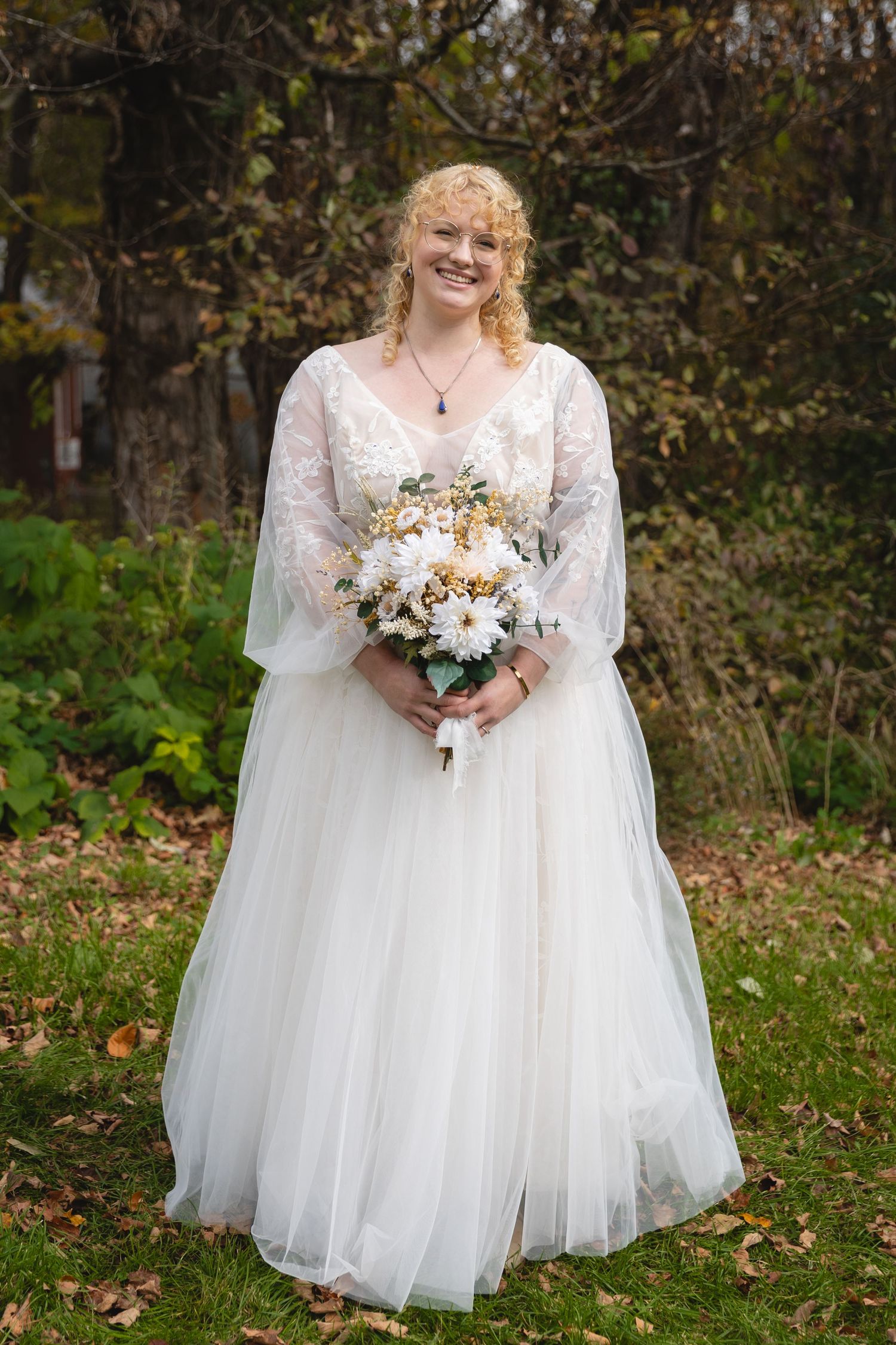 Bride in flowing white dress holds bouquet while standing in autumn garden.