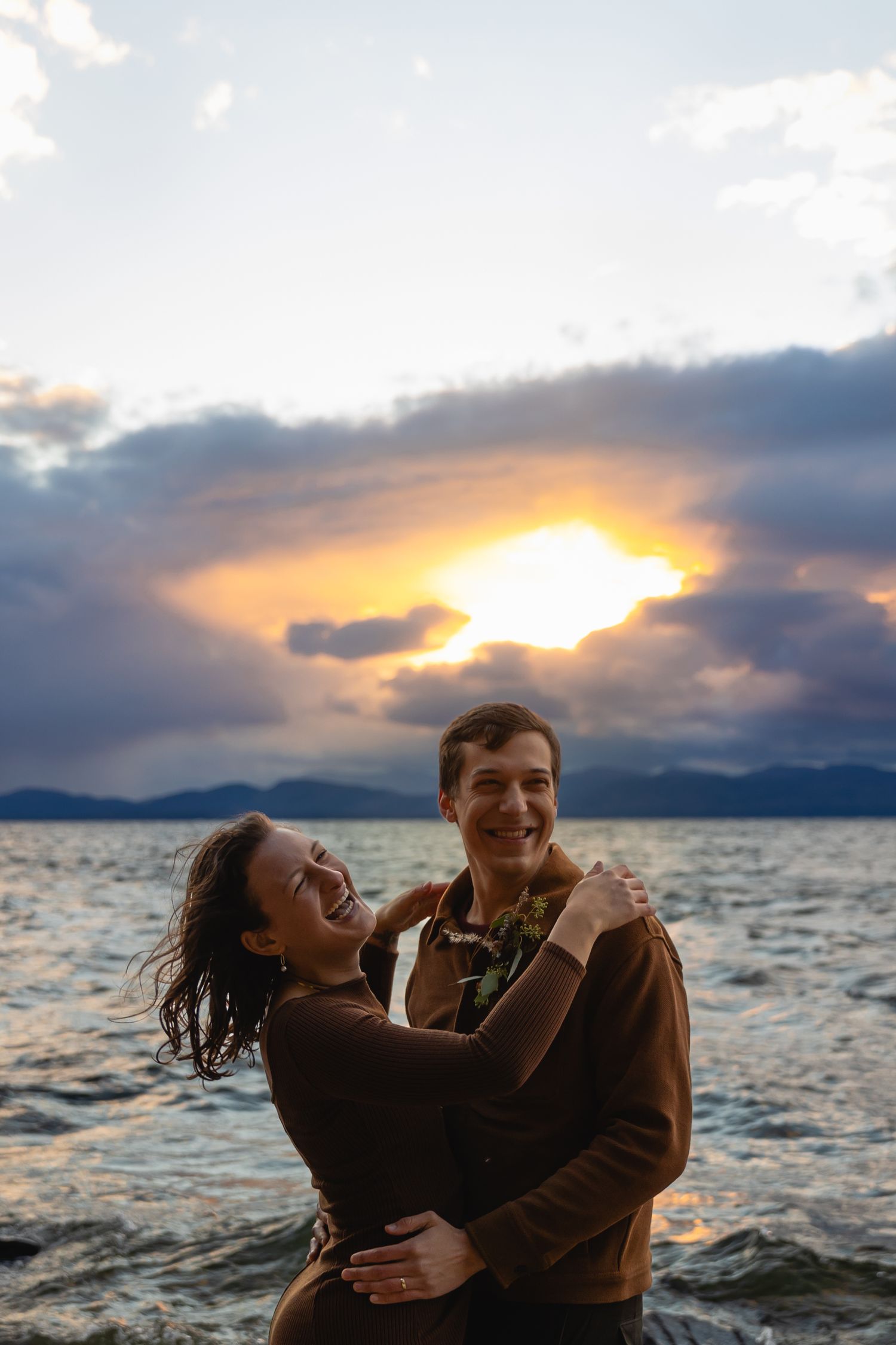 A couple embraces joyfully by a lake at sunset with dramatic clouds and mountains in the background.