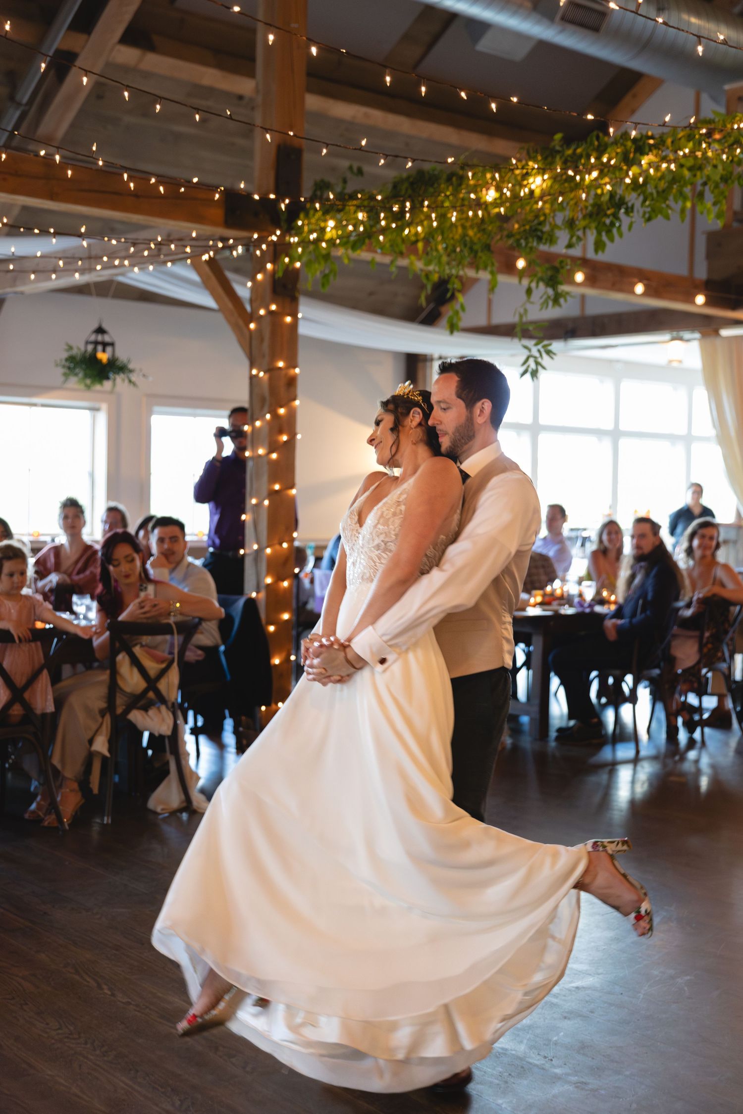 Couple performing dance moves in rustic reception venue with string lights and wooden beams.