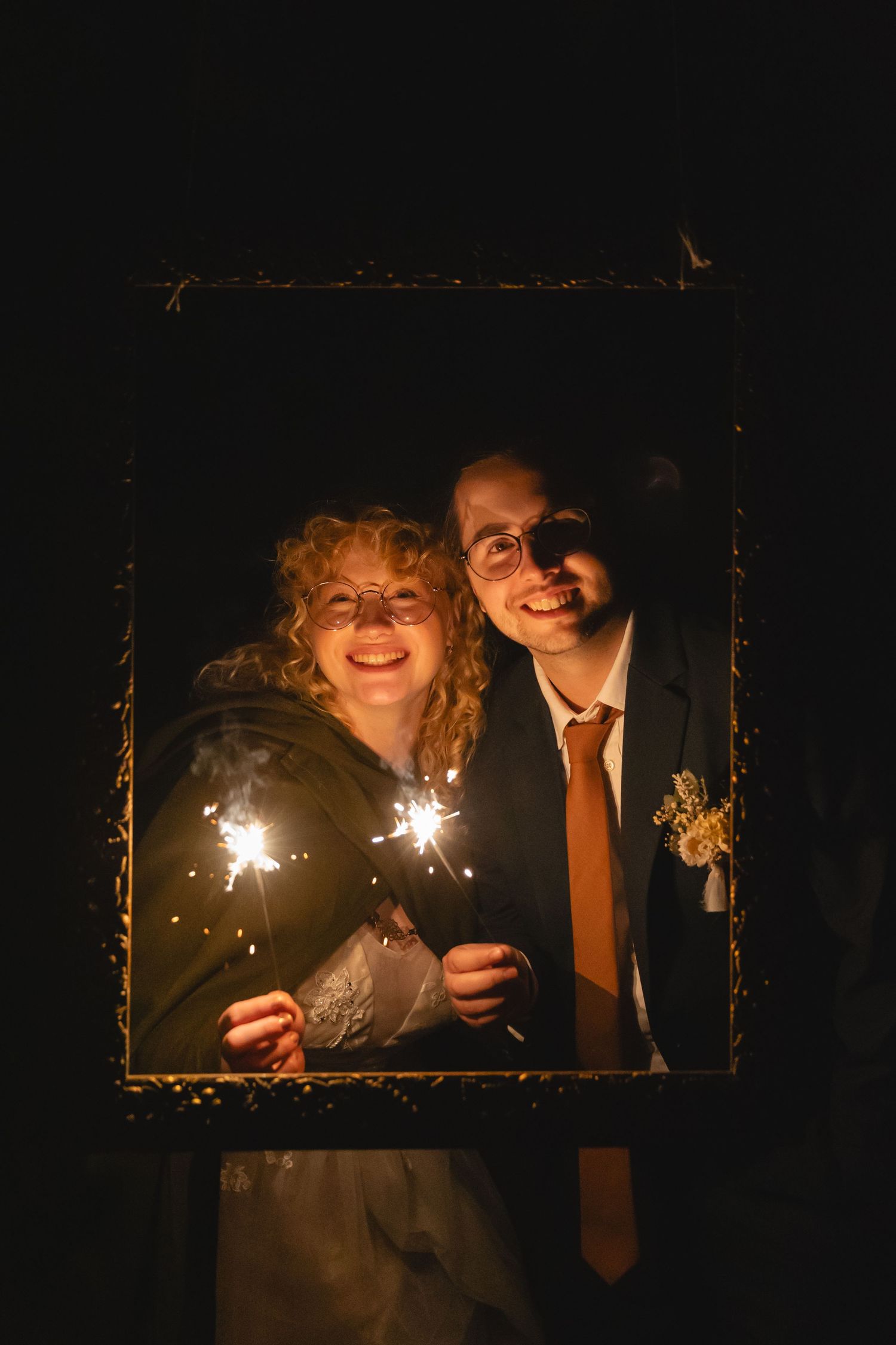 Couple holds sparklers while framed in a decorative border at night.