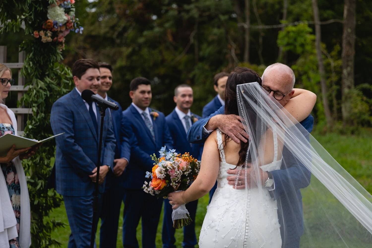 Wedding party looks on as couple shares emotional moment during outdoor ceremony with flowing veil and peach bouquet.