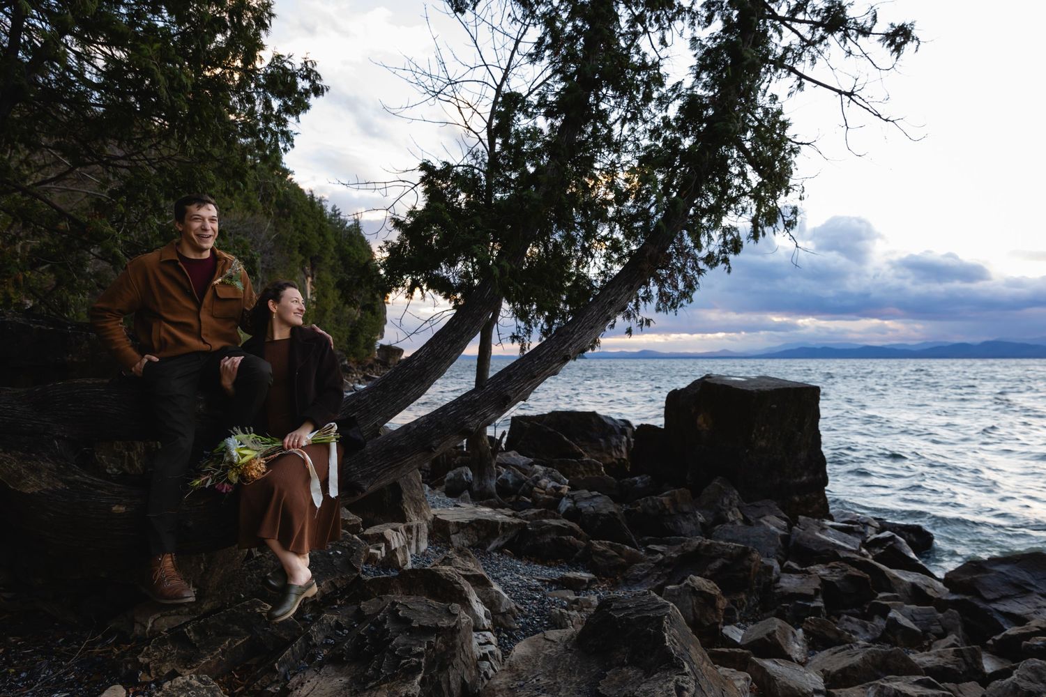 Two people sit on rocky shoreline beneath leaning tree with dramatic cloudy skies and lake view in background.