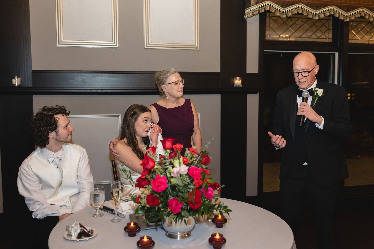 People sitting at a round table with red rose centerpiece laugh while someone speaks at a formal event.