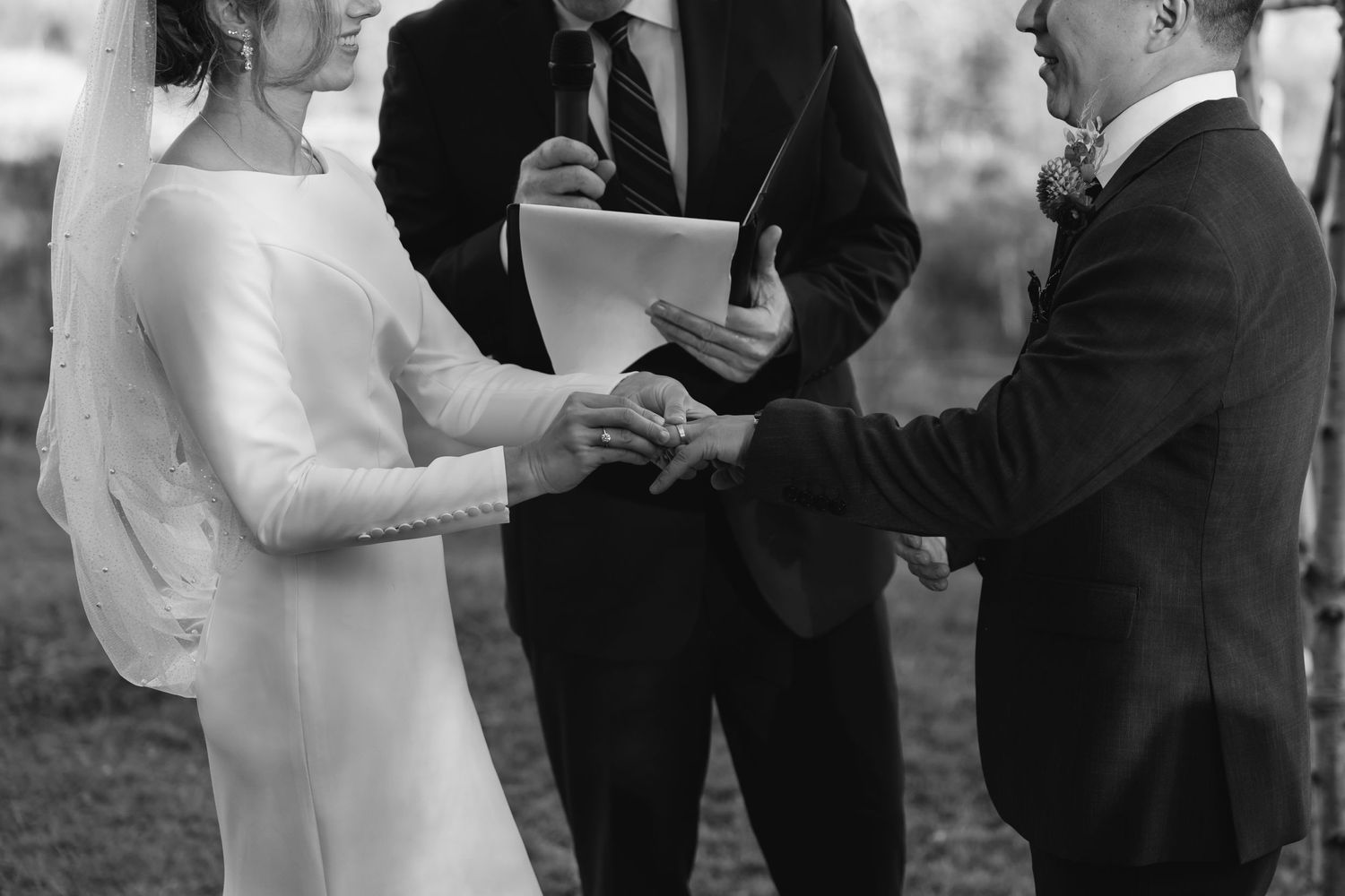 A black and white wedding ceremony photograph shows a couple holding hands while officiant leads their vows outdoors.