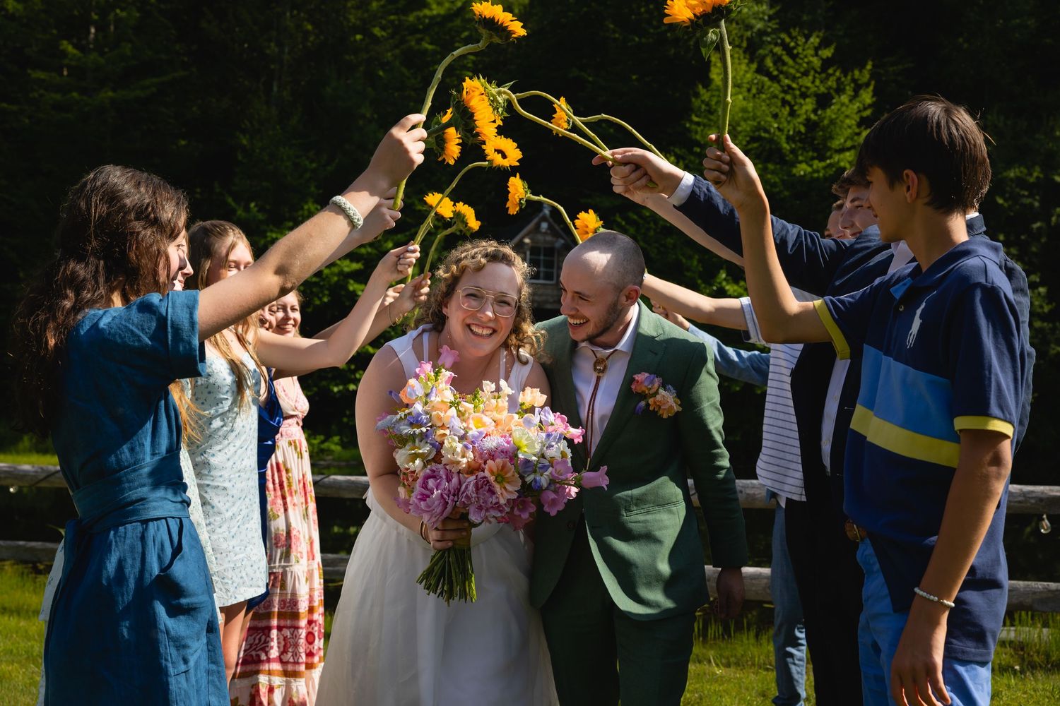 Wedding party celebrates with raised arms while walking in a line formation through a sunny garden setting.